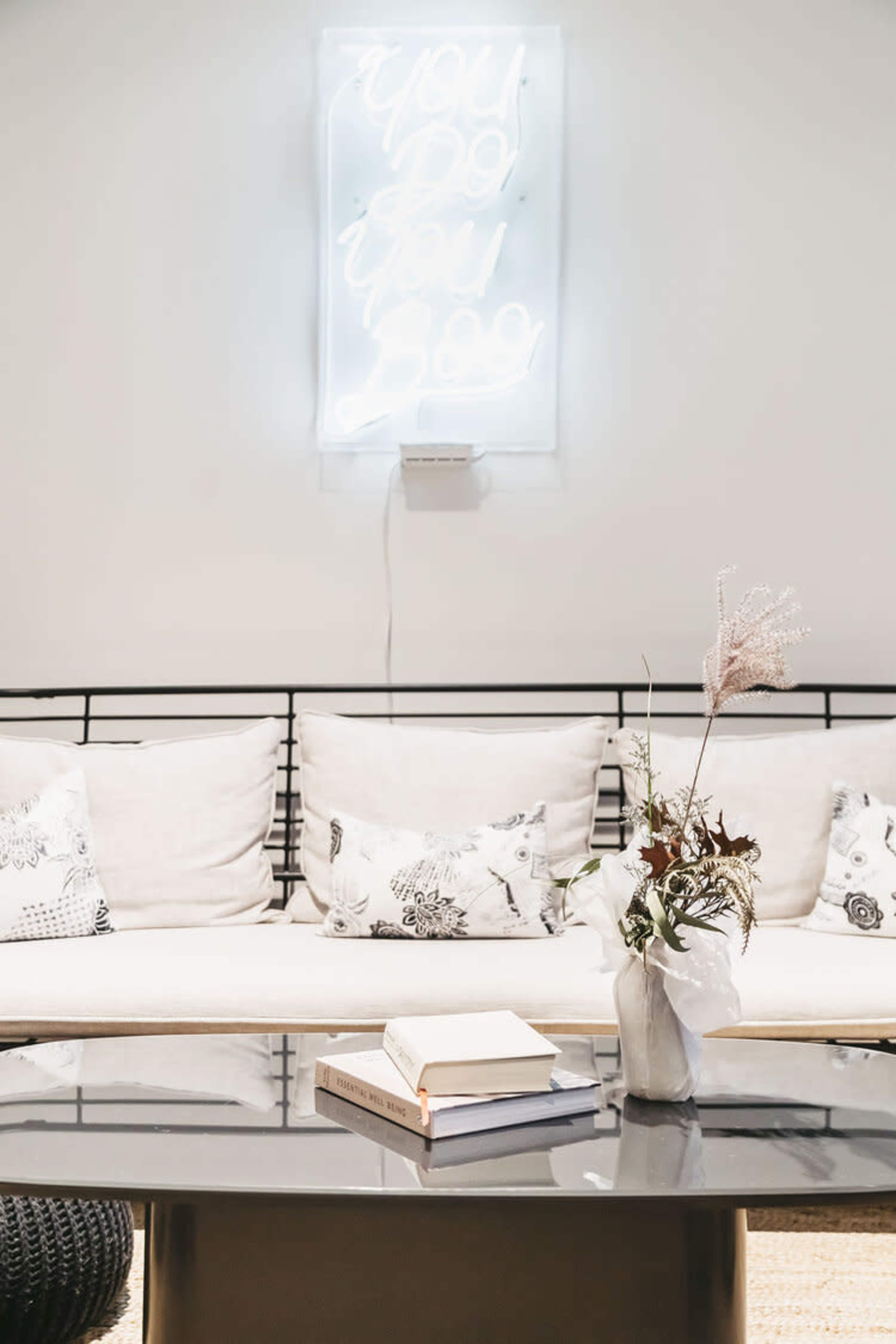 A minimalist living space features a neon sign, a white sofa with decorative pillows, a coffee table with stacked books, and a vase of dried flowers.