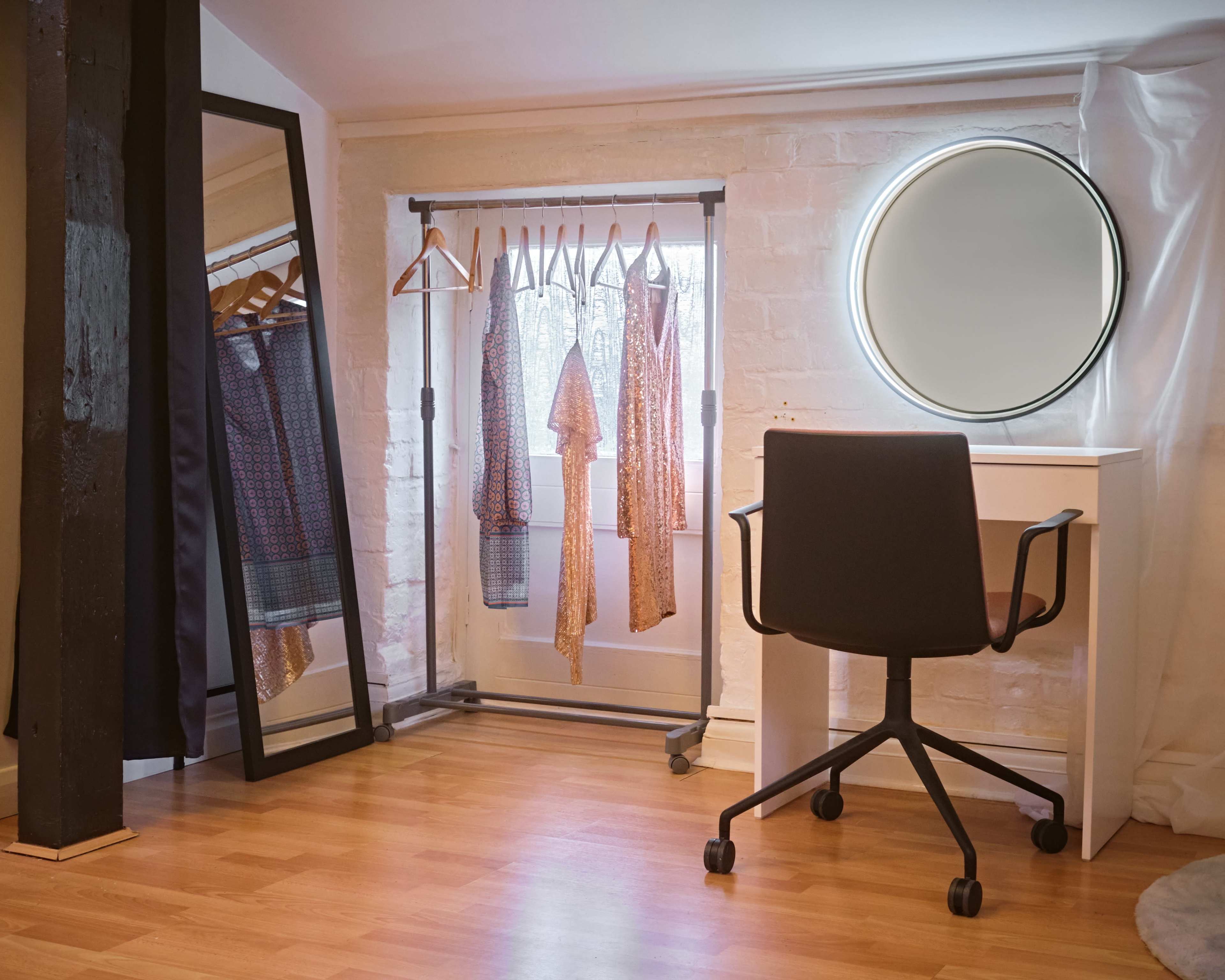 A minimalist dressing area features a clothing rack with several garments, a round mirror, and a black wheeled chair against a backdrop of light-colored walls and wooden flooring.