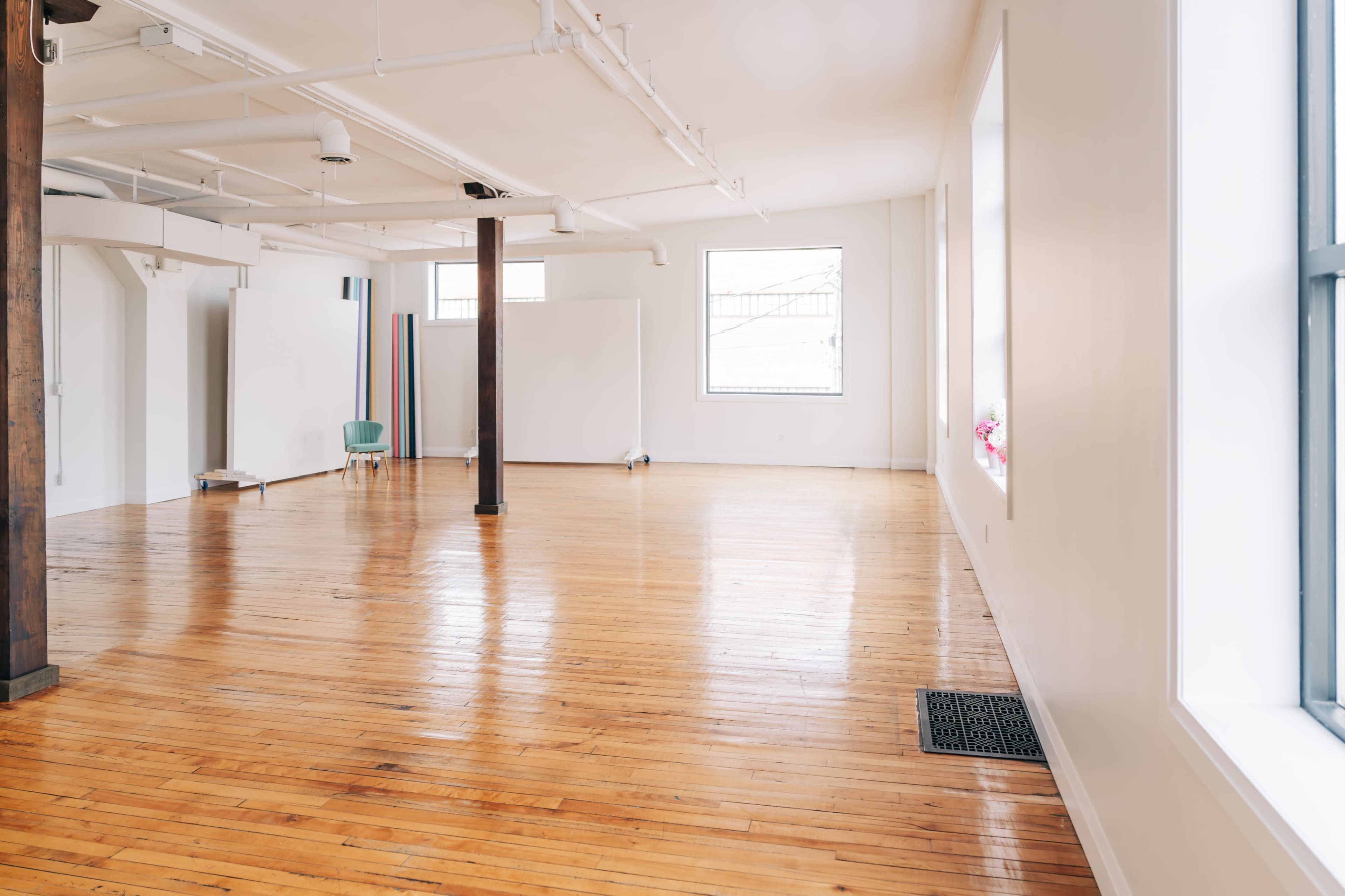 A spacious, empty room with hardwood floors, large windows, and a few pieces of minimalist furniture against the walls.