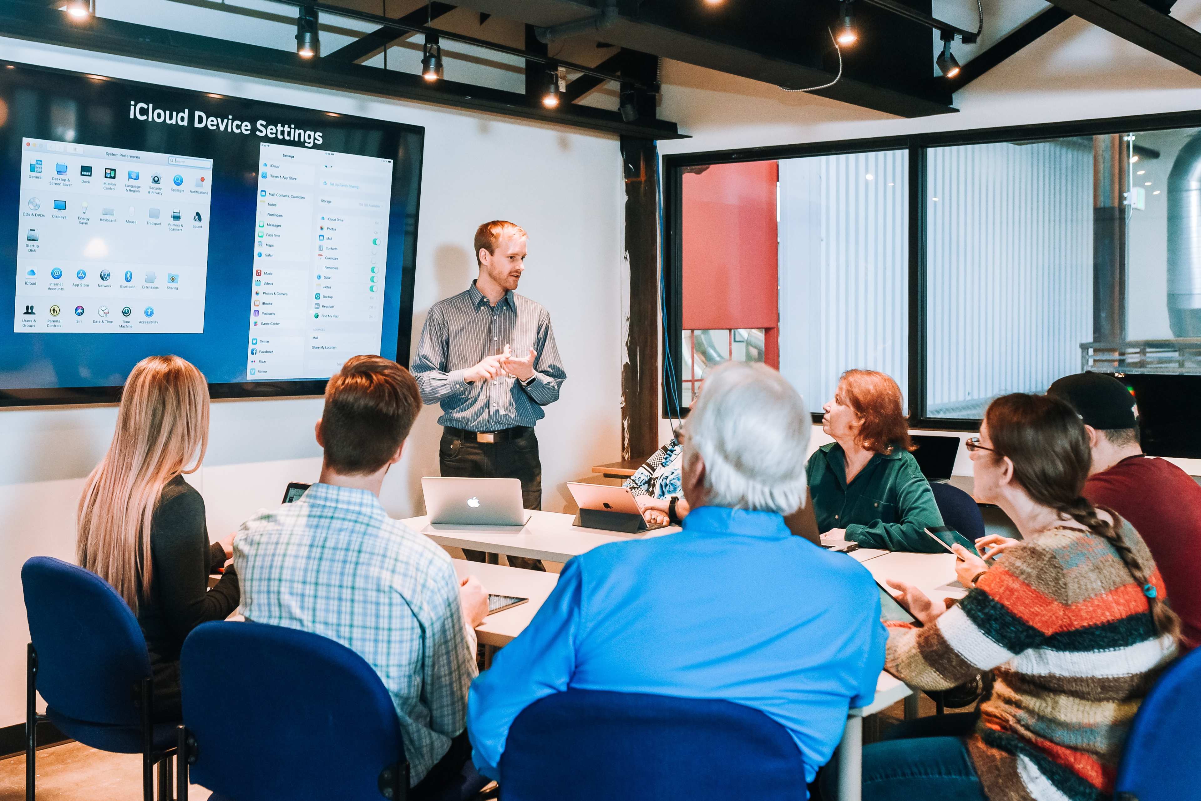 A presenter stands in front of a group of seated individuals in a conference room, engaging them while a large screen displays iCloud device settings.
