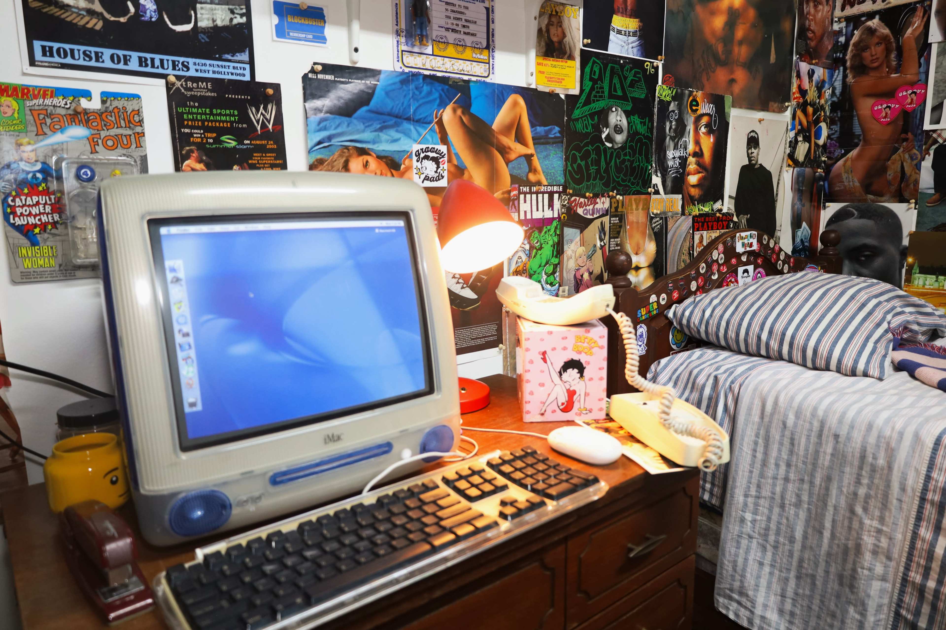 The image shows a vintage computer on a wooden desk beside a landline phone, with a cluttered wall covered in posters and various memorabilia, and a bed in the background.