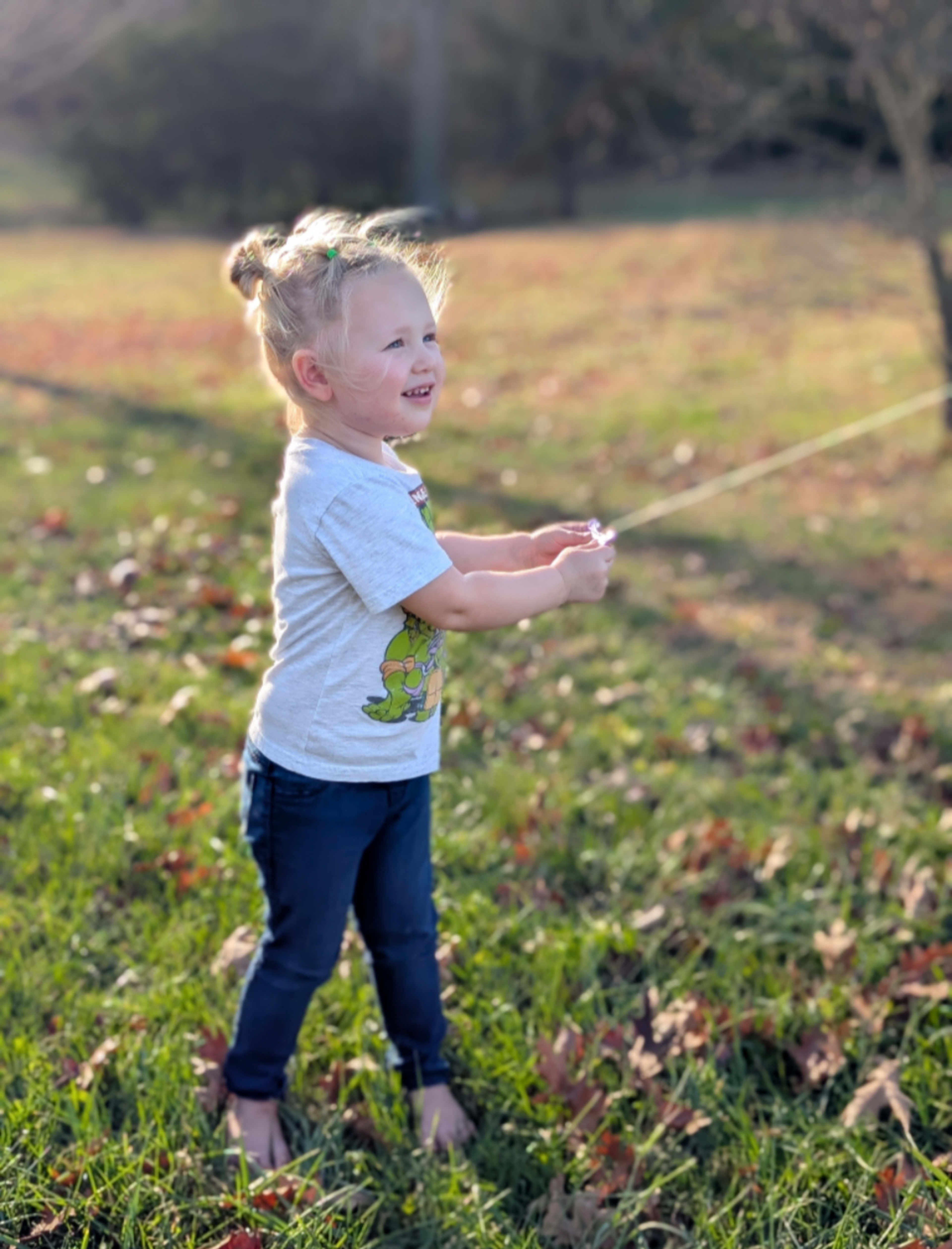 A young child stands in a grassy field, holding a string and looking up with a smile.