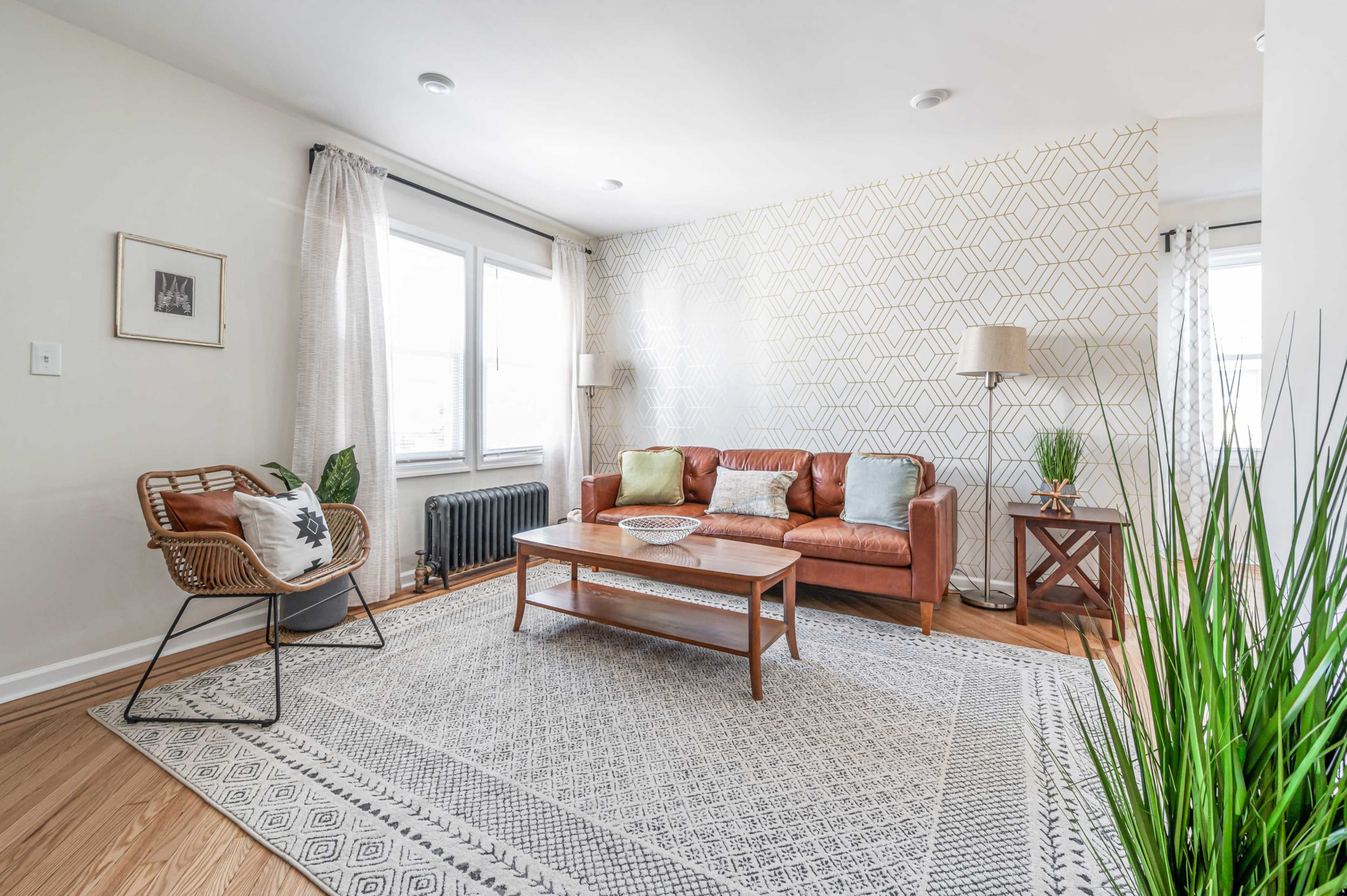 A bright living room featuring a leather sofa, a patterned rug, a wooden coffee table, and a large window with sheer curtains.