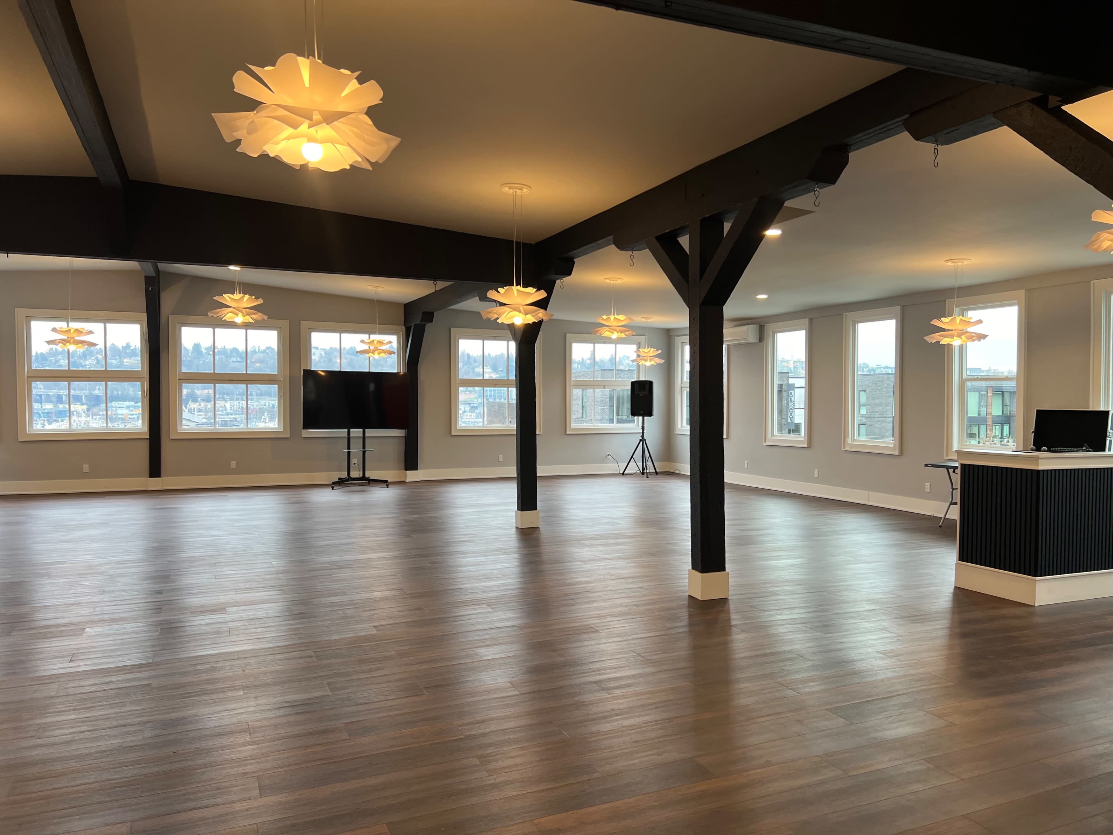 An empty event space with wooden flooring, large windows, and modern light fixtures hanging from the ceiling.