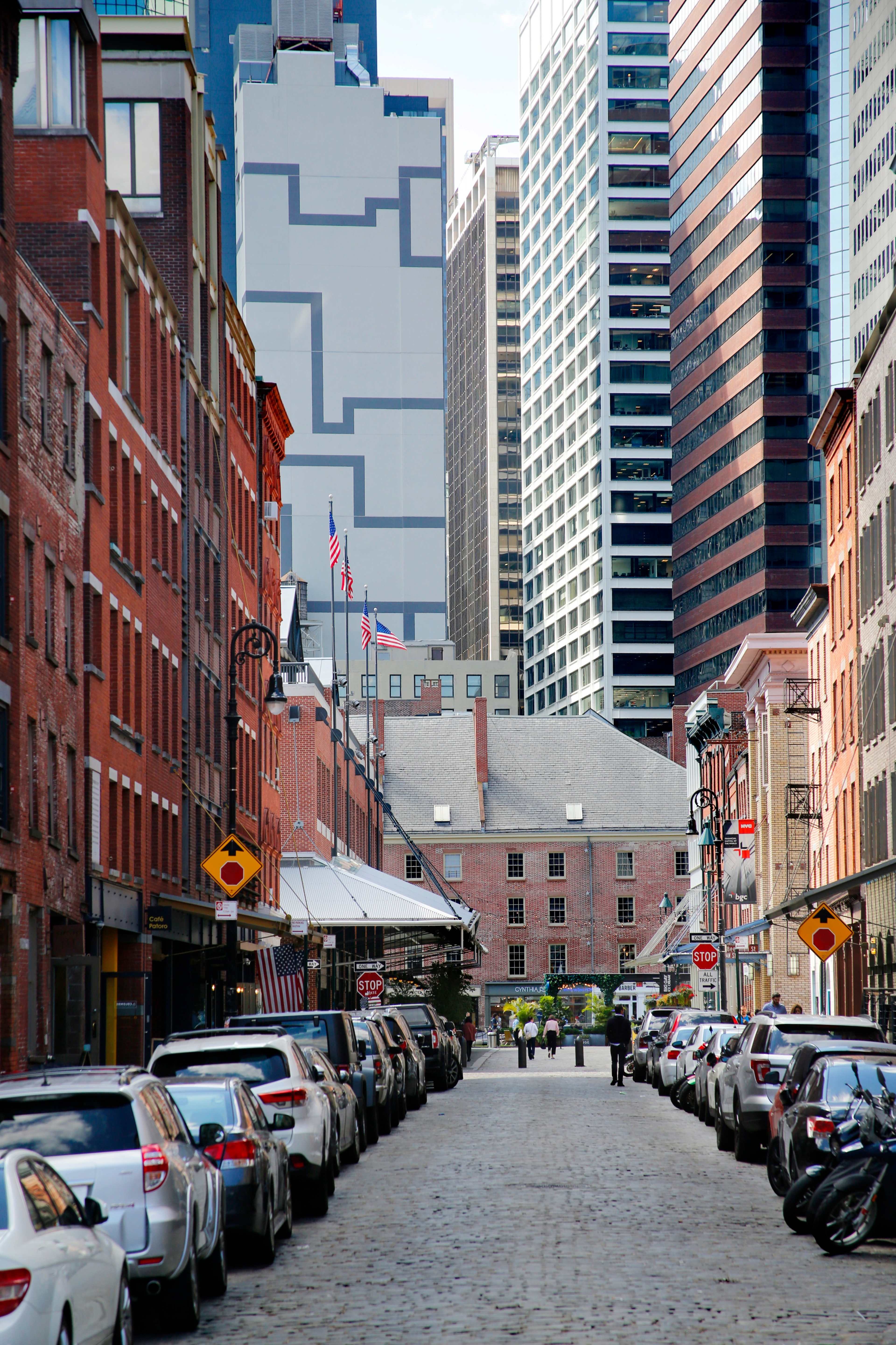 A cobblestone street lined with parked cars leads to a view of tall modern buildings and a historic brick structure.