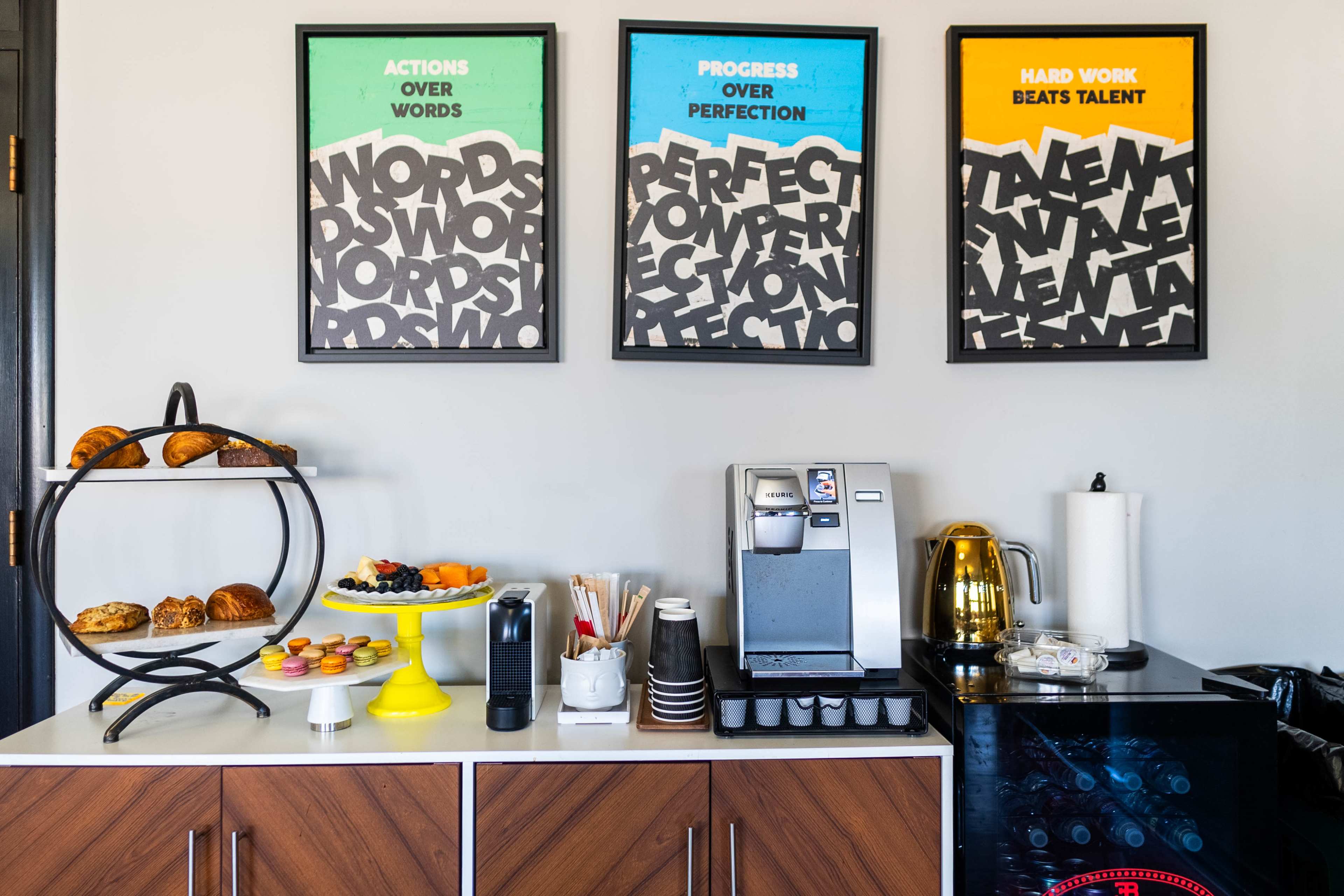 The image shows a refreshment station featuring a tiered stand with pastries, a coffee machine, a yellow cake stand with dessert items, and three framed motivational posters on the wall above.