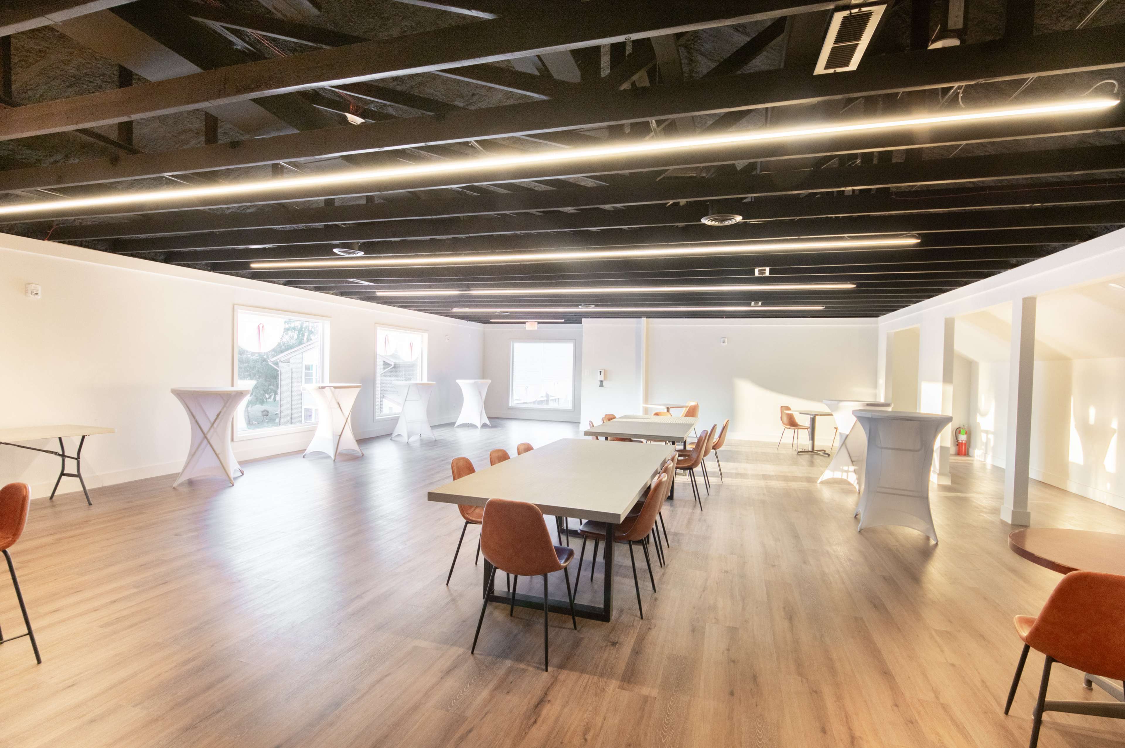 A spacious room with wooden flooring, multiple tables and chairs, and modern lighting.