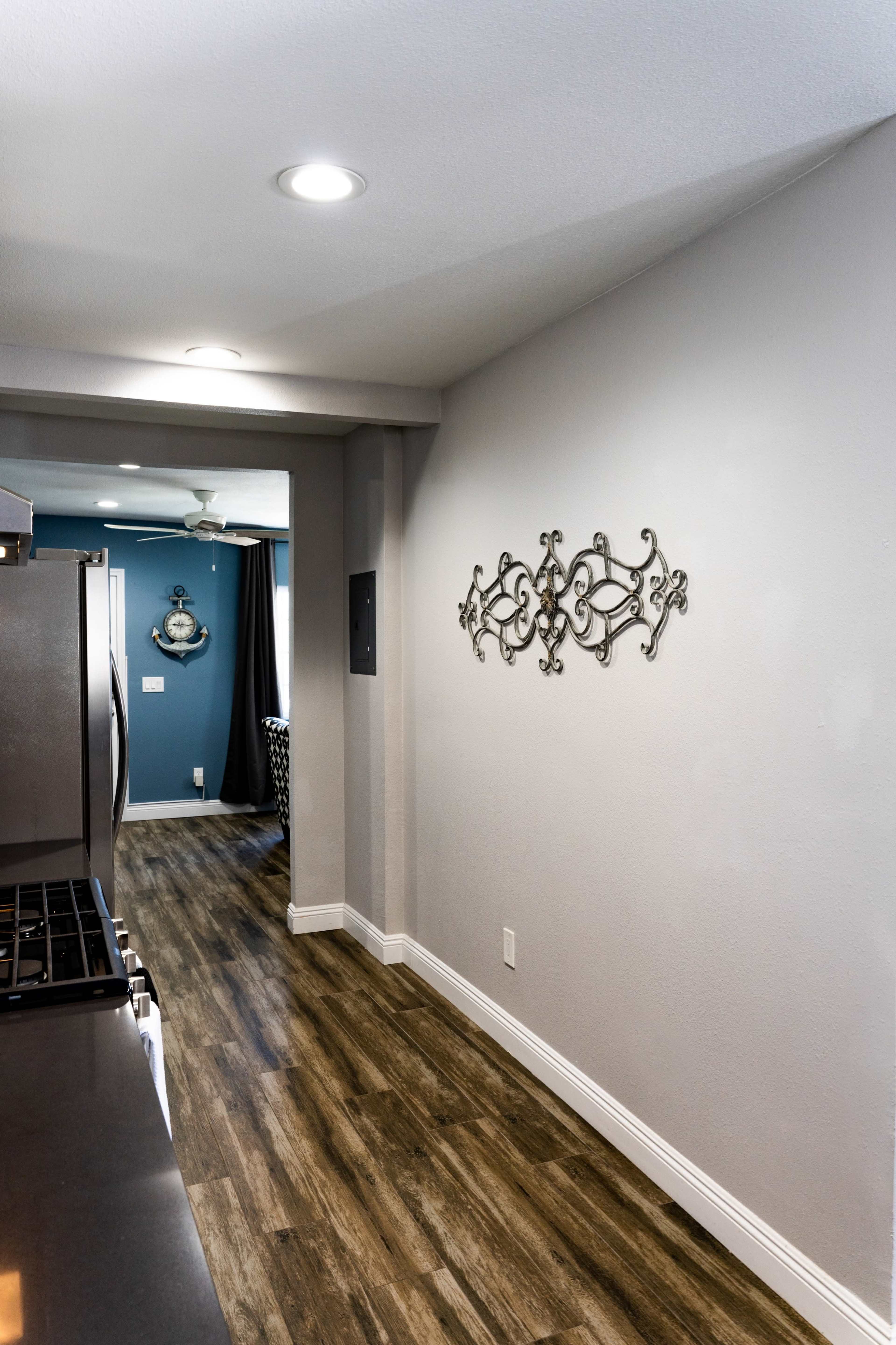 The image shows a narrow kitchen hallway with dark wood-like flooring, a stainless steel refrigerator on the left, and decorative wall accents on the right, leading to an open living area.