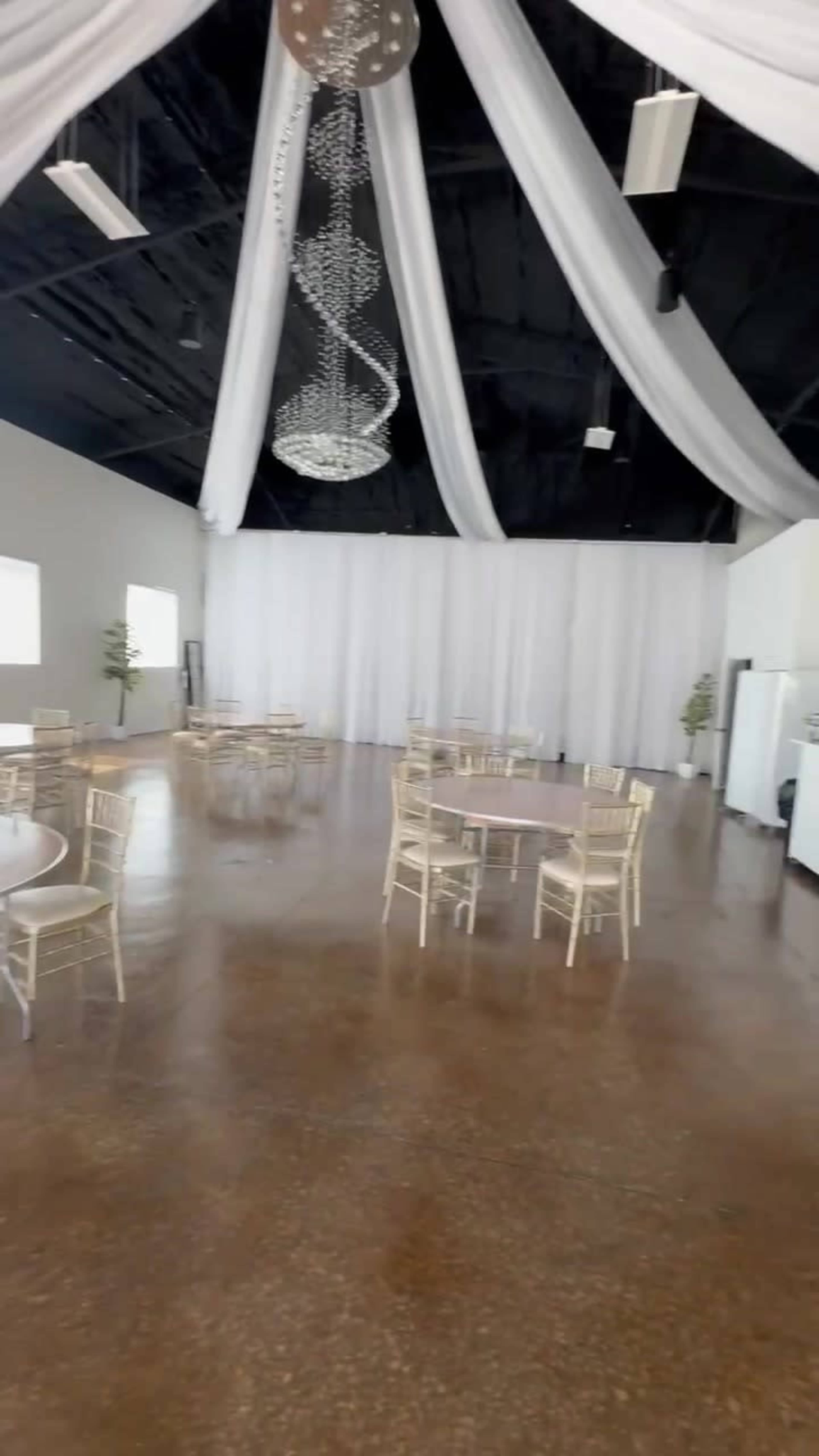 The image shows an empty event space featuring several round tables, chairs, and a draped ceiling with a chandelier.