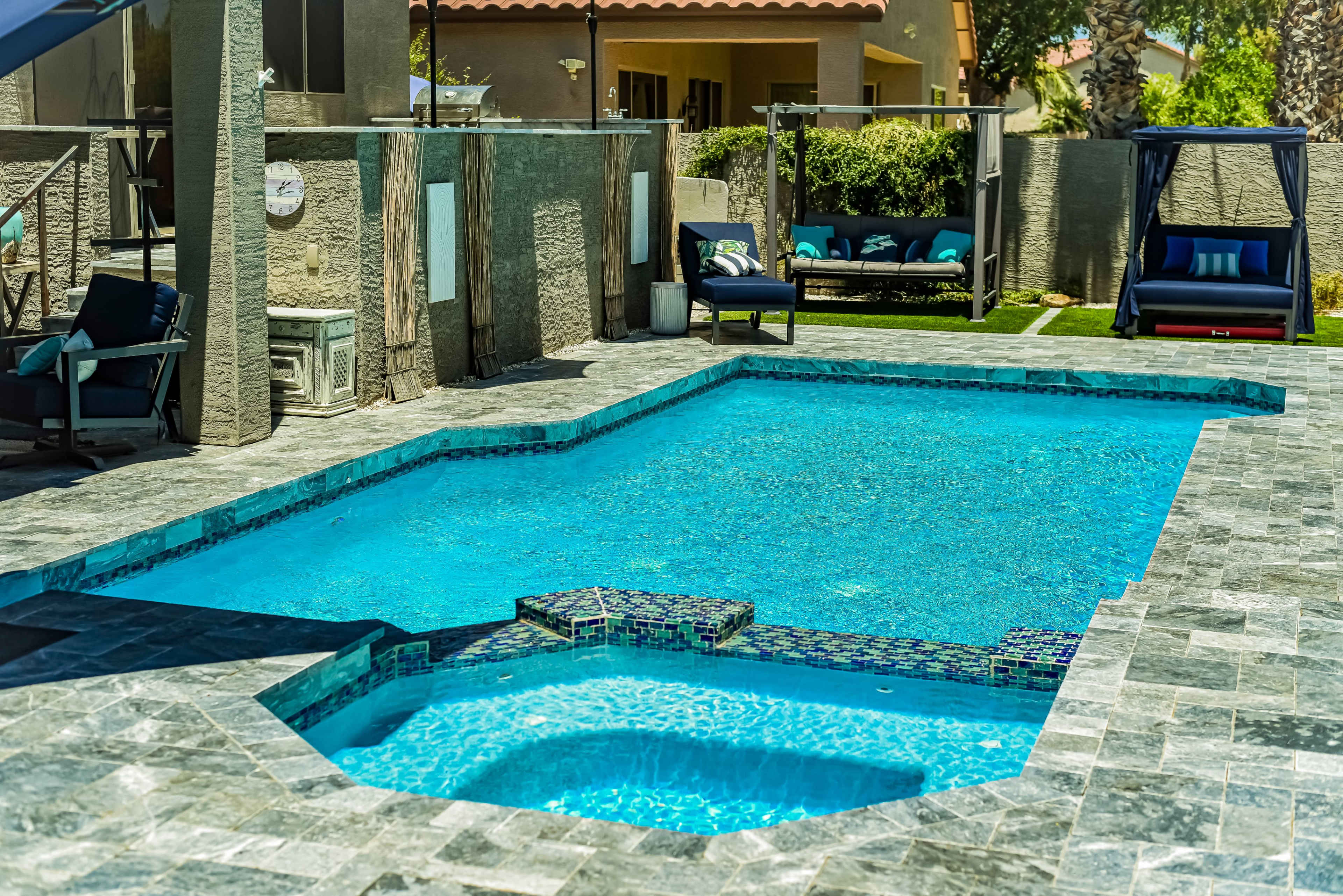 A rectangular swimming pool surrounded by gray stone patio tiles and lounge chairs in a backyard setting.