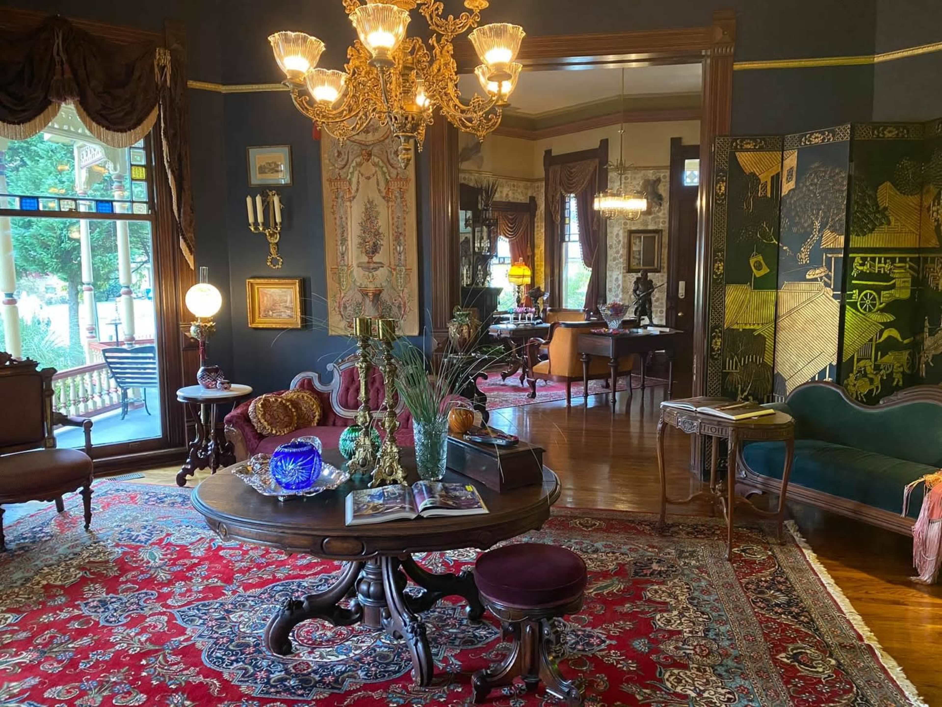 The image shows an elegantly decorated parlor featuring a vintage chandelier, a round table with decorative items, a richly patterned rug, and richly adorned walls.