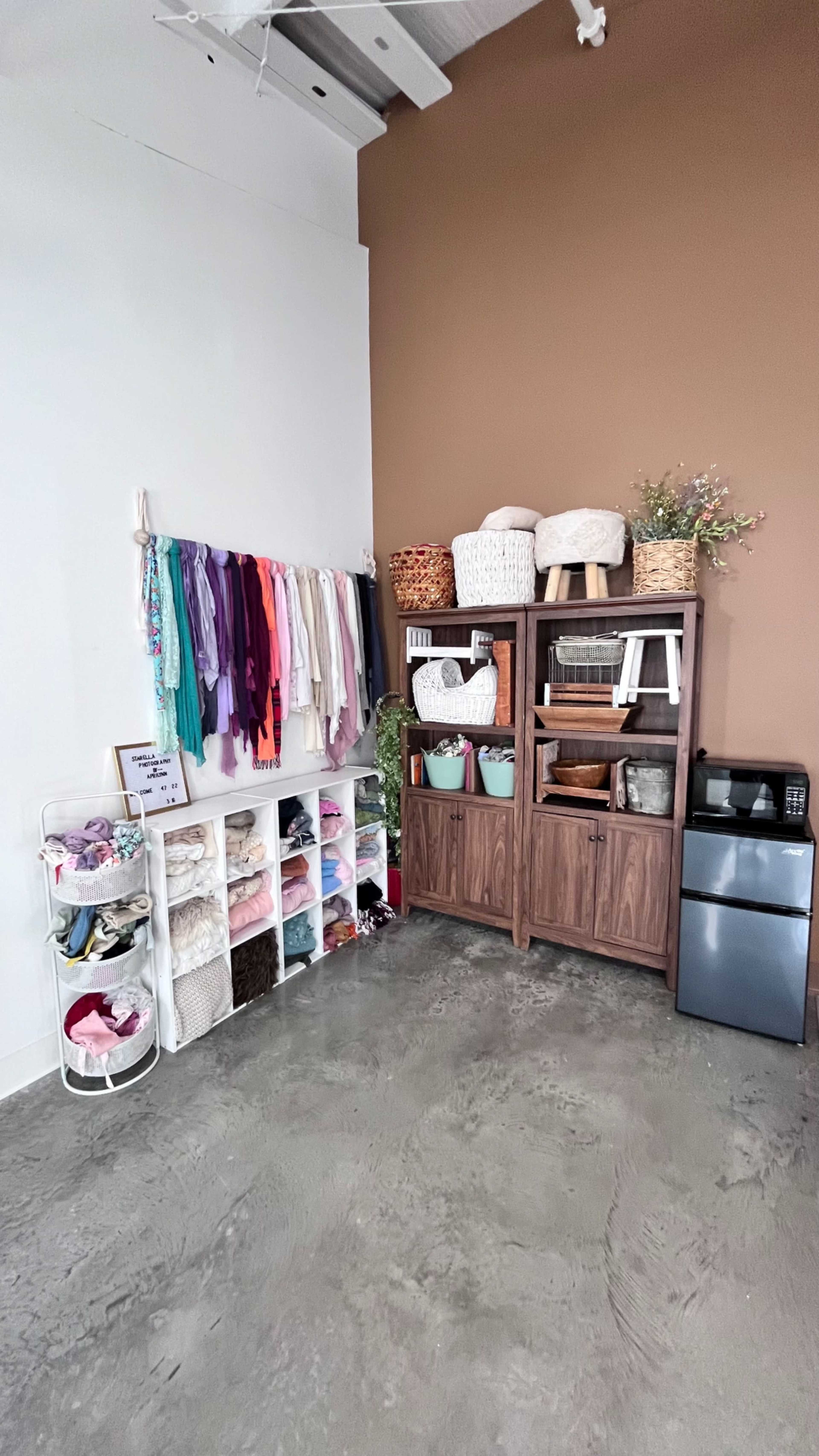 The image shows a neatly organized corner of a room with shelves displaying various textiles and containers, along with a small refrigerator.