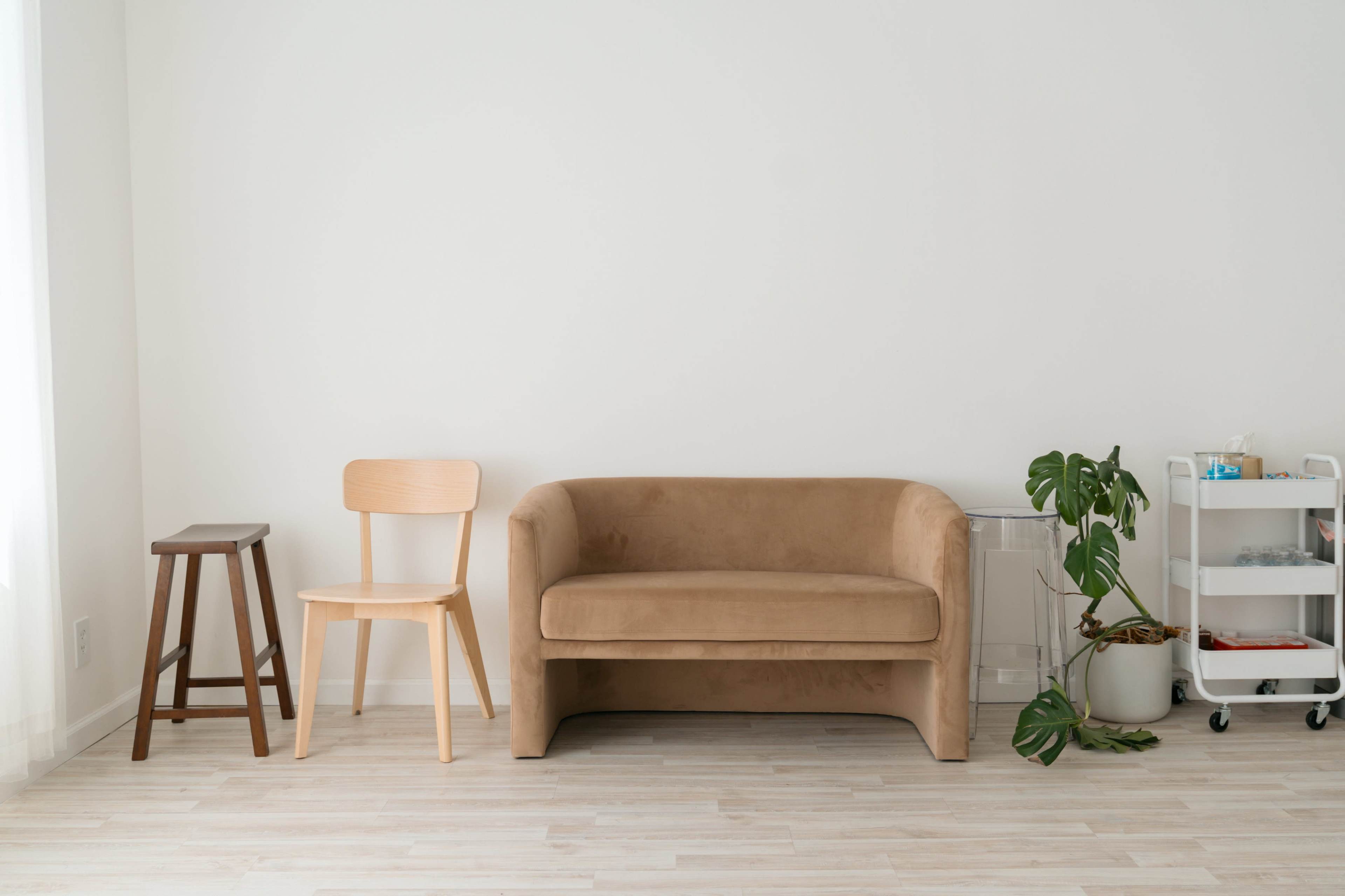 The image shows a simple living space with a brown sofa, a light wooden chair, a small wooden stool, and a cart holding various items beside a plant against a white wall.