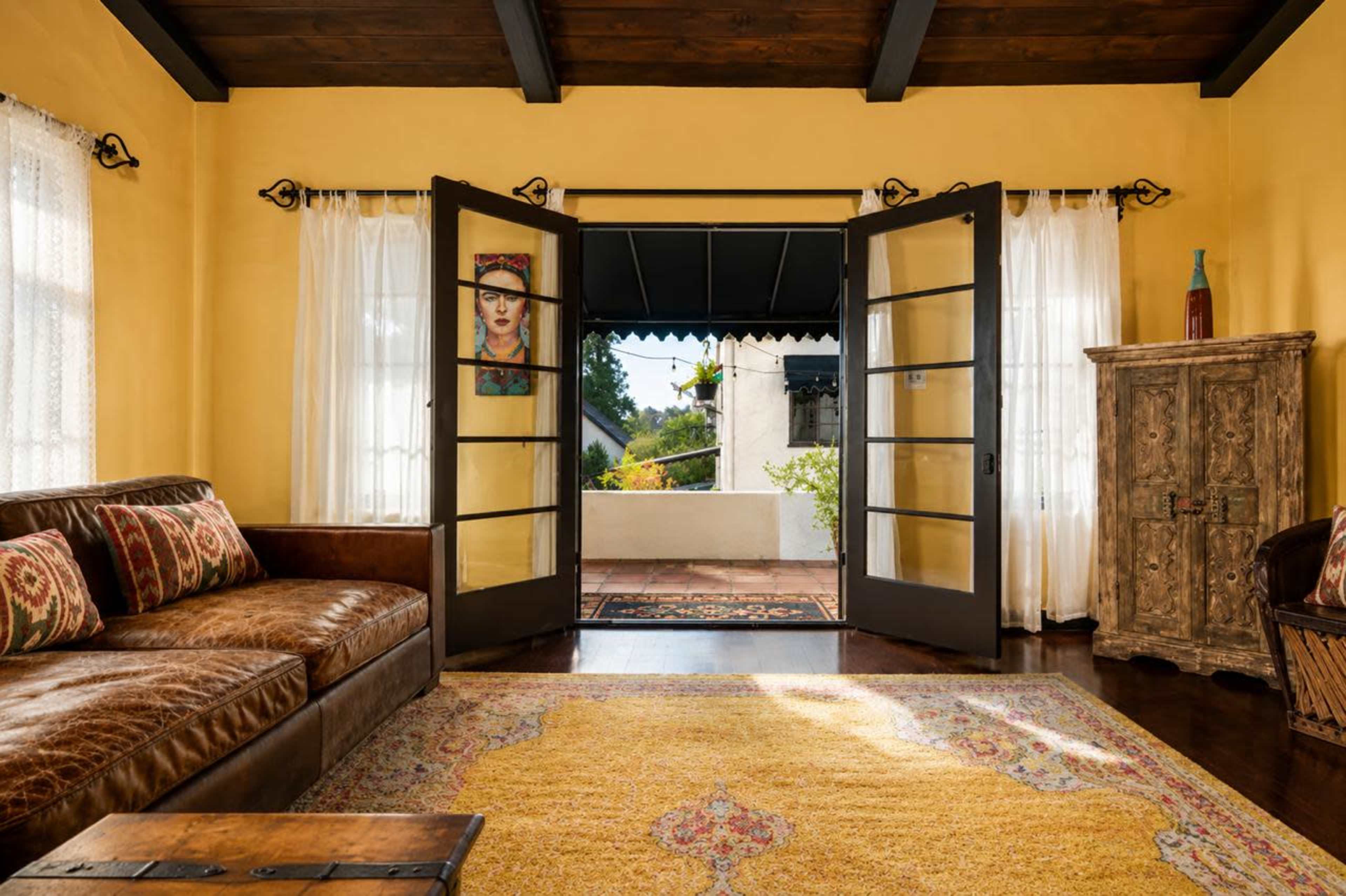 A cozy interior features a leather sofa, large glass doors opening to a patio, and warm yellow walls adorned with curtains and a piece of art.