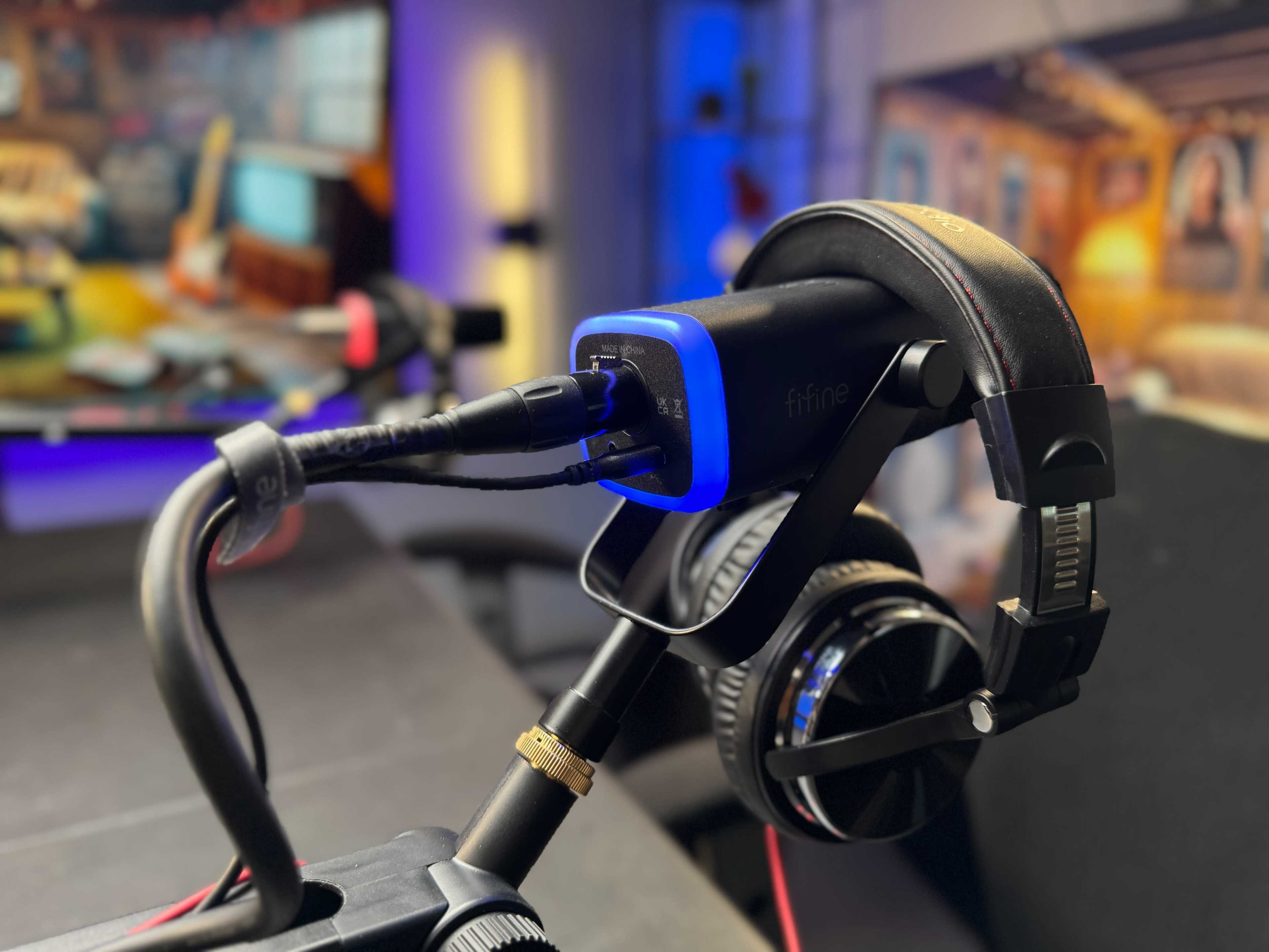 A microphone and headphones are positioned on a stand in front of a blurred background featuring lighting and equipment typically found in a studio setting.