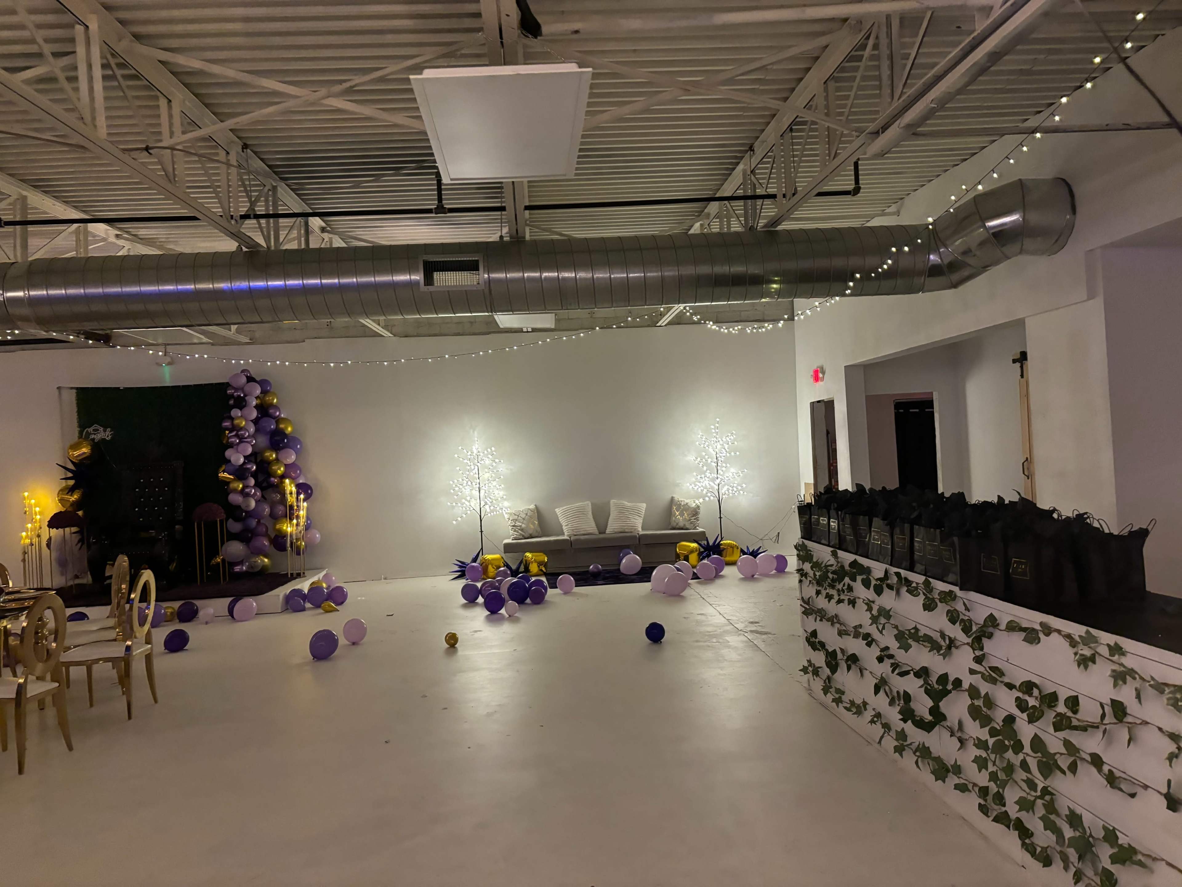 The image shows a decorated event space featuring purple and gold balloons, two lighted trees, and a seating area with a white sofa against white walls.