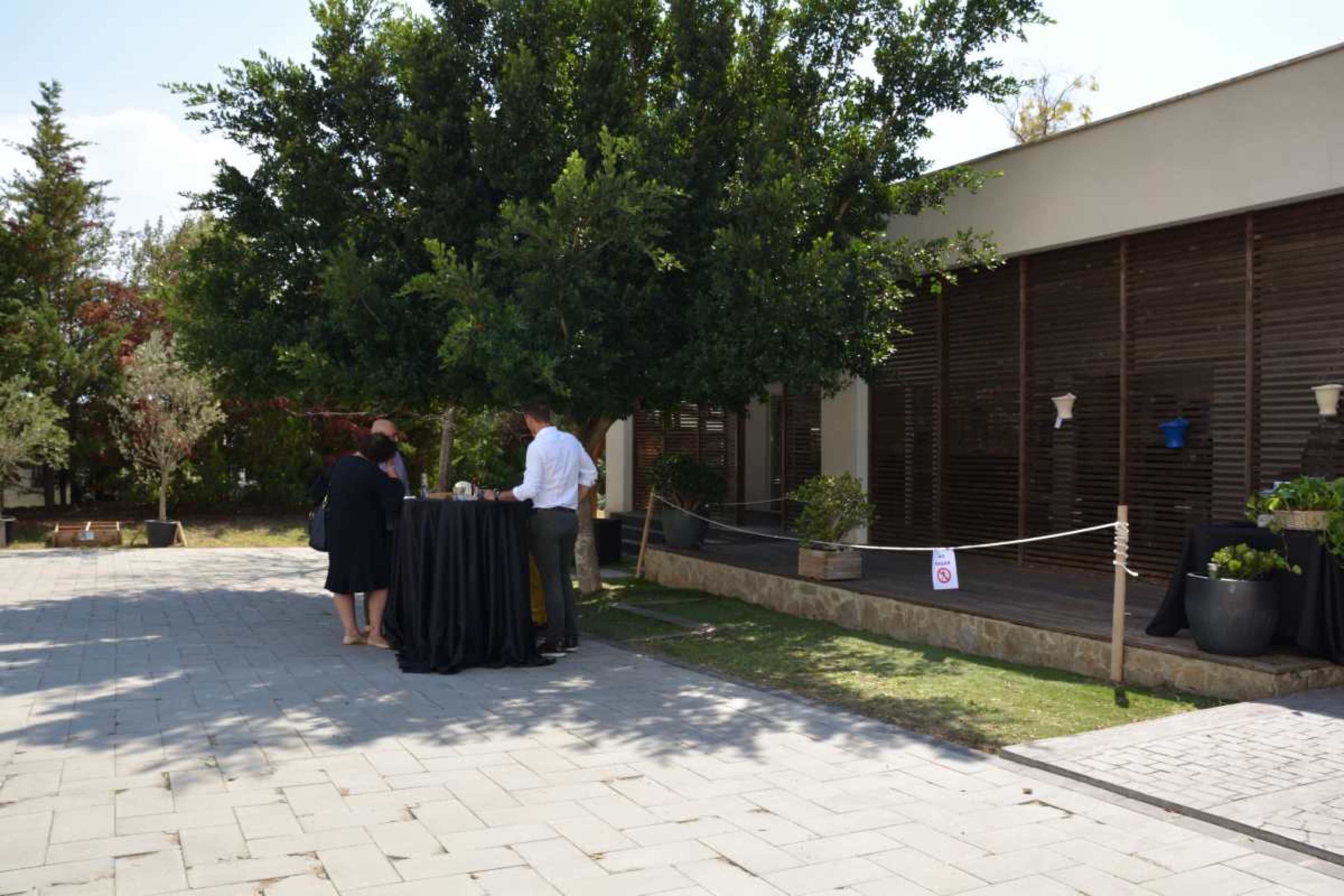 A couple stands near a table under a large tree in a landscaped outdoor area adjacent to a modern building.