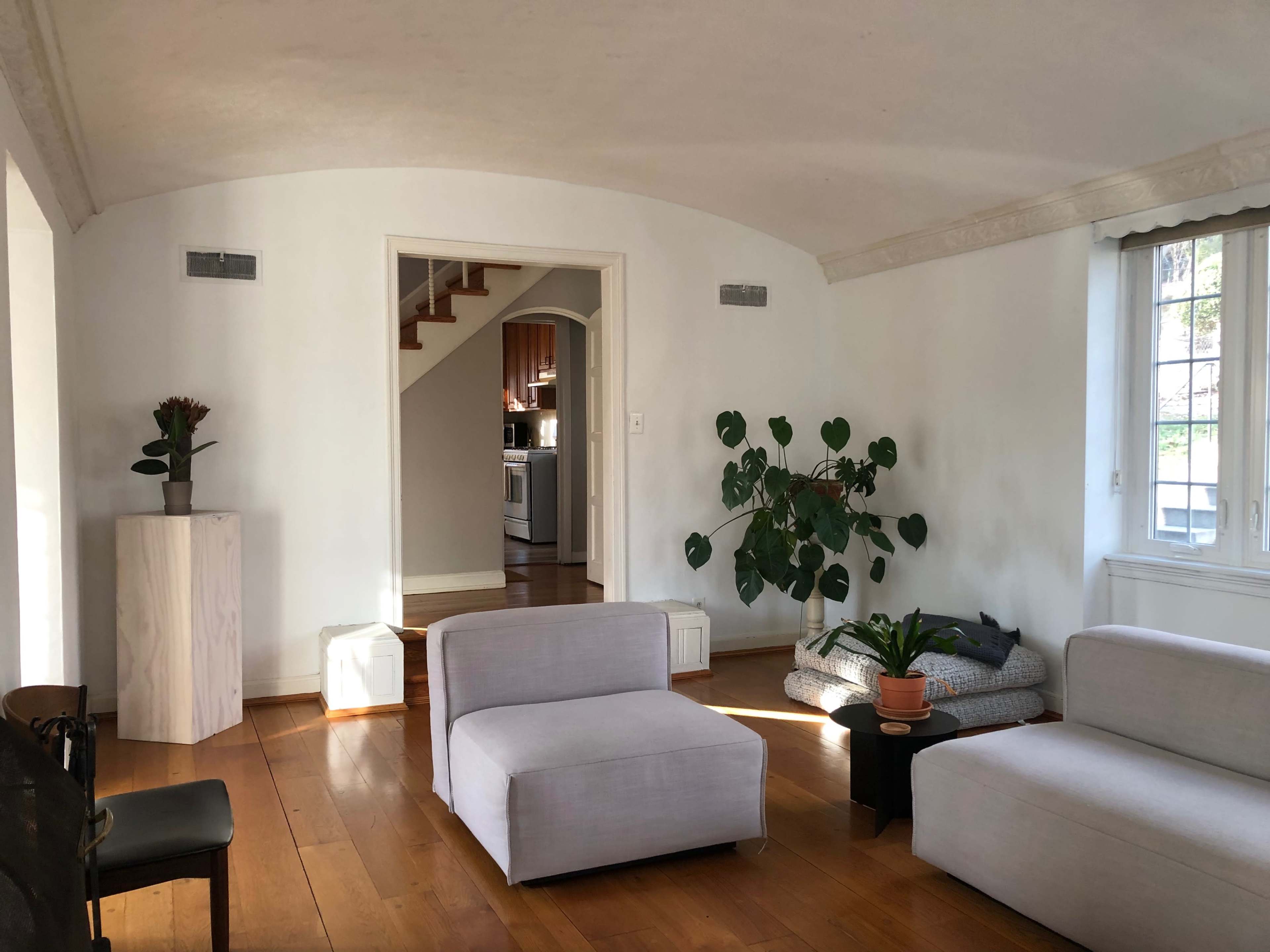A modern living room with light-colored walls, wooden flooring, and a large window illuminating a few pieces of furniture, including a plant and a staircase leading to another area.