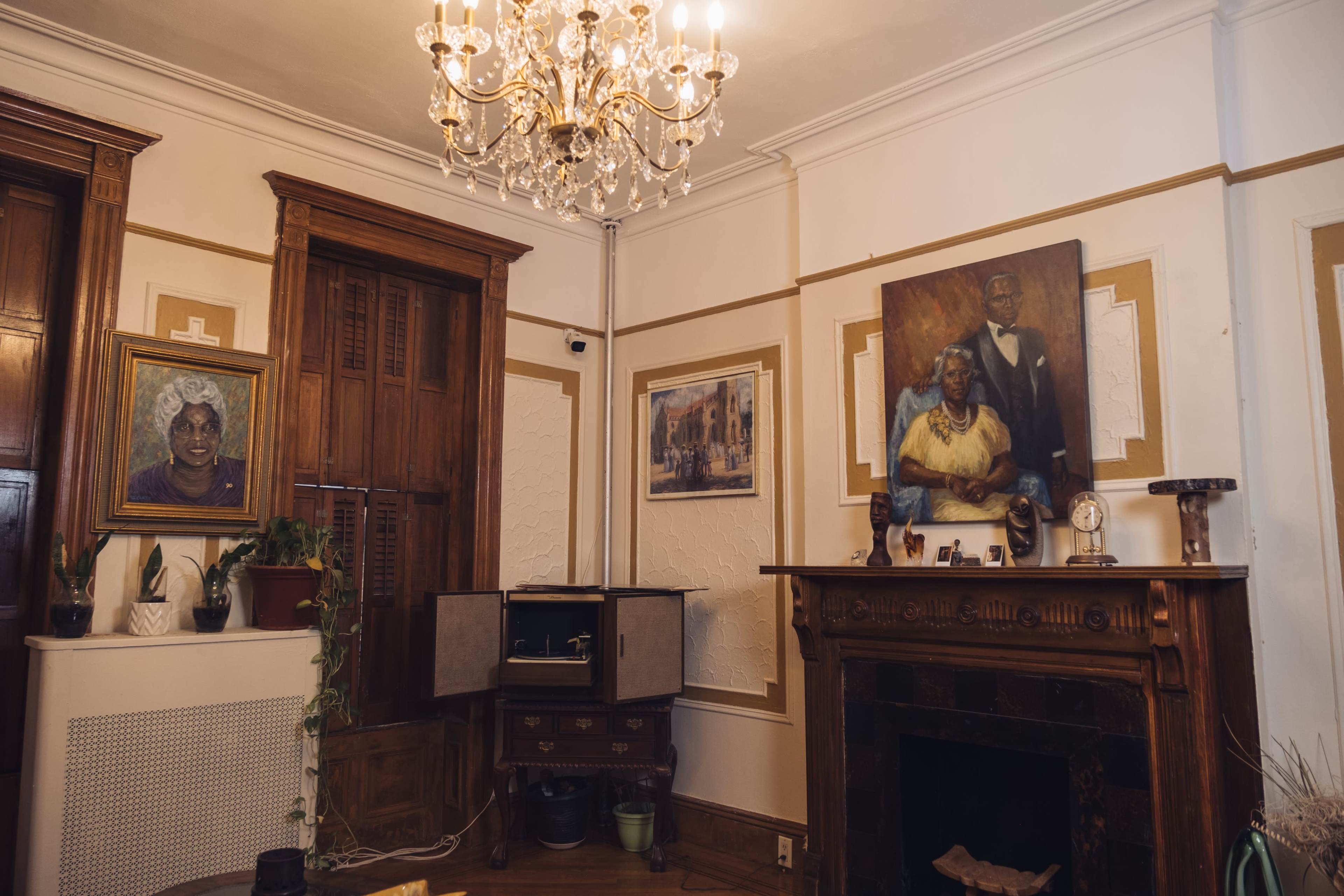 The room features a vintage fireplace, a chandelier, and framed paintings on the walls, alongside an antique record player.
