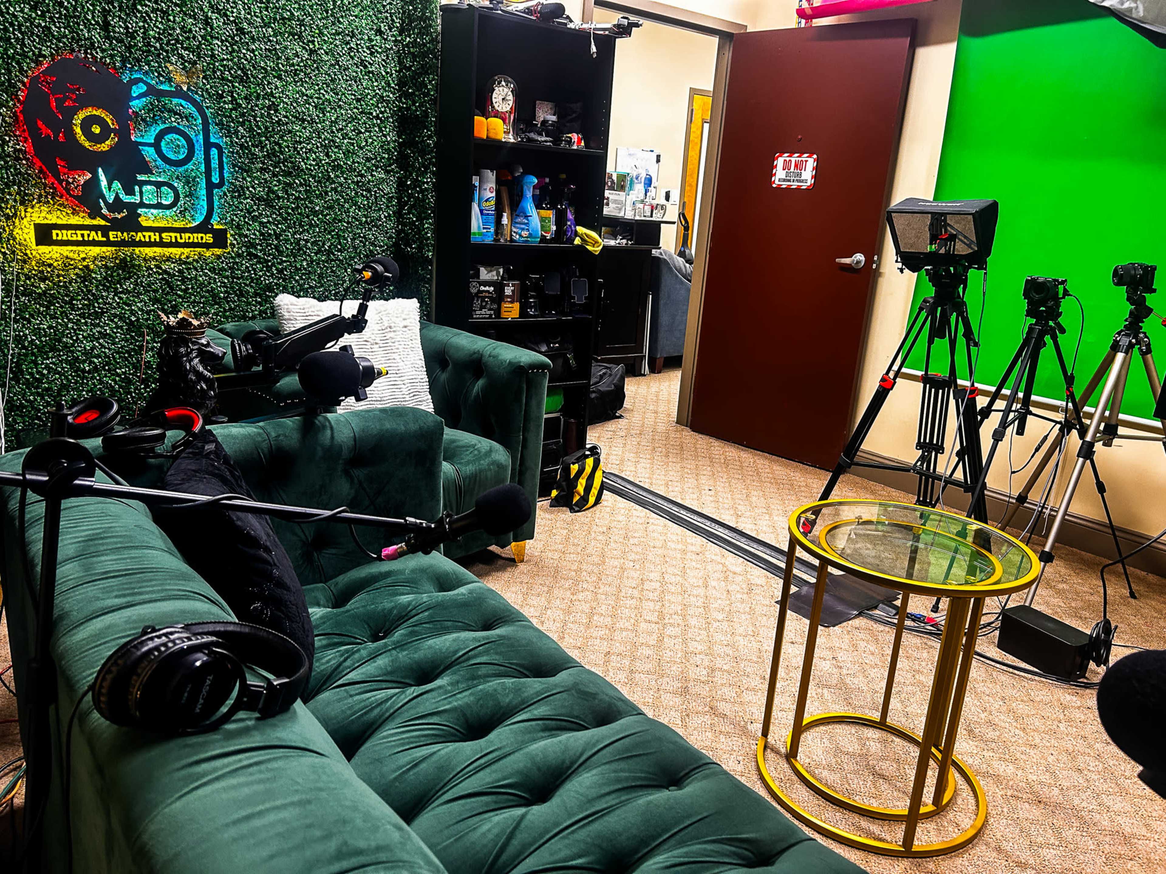 The image shows a podcast studio with a green wall, two cameras on tripods, a small gold coffee table, and a green velvet couch with microphones nearby.