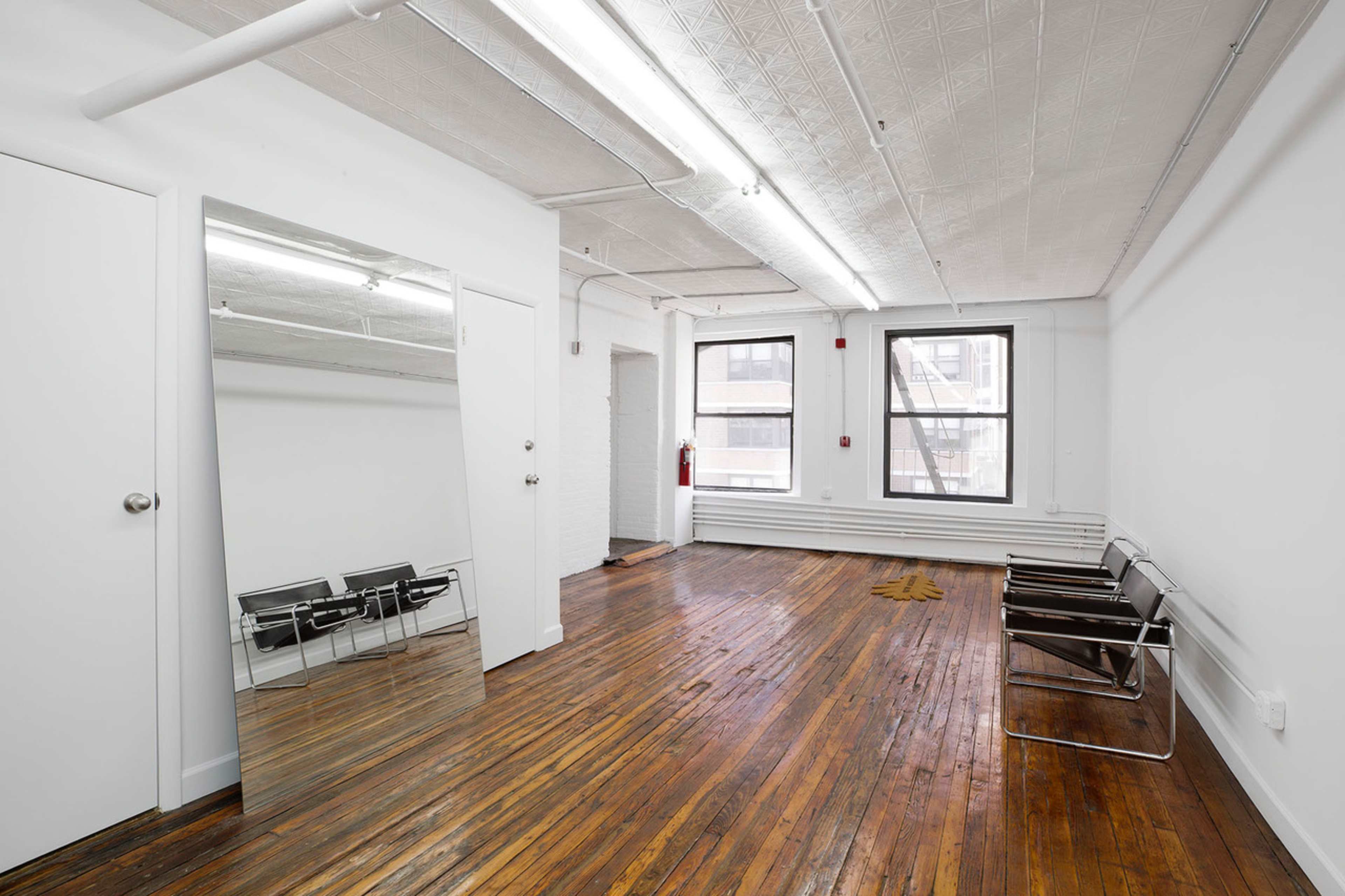 The image shows a spacious, empty room with hardwood floors, two large windows, a tall mirror leaning against the wall, and a pair of black chairs.