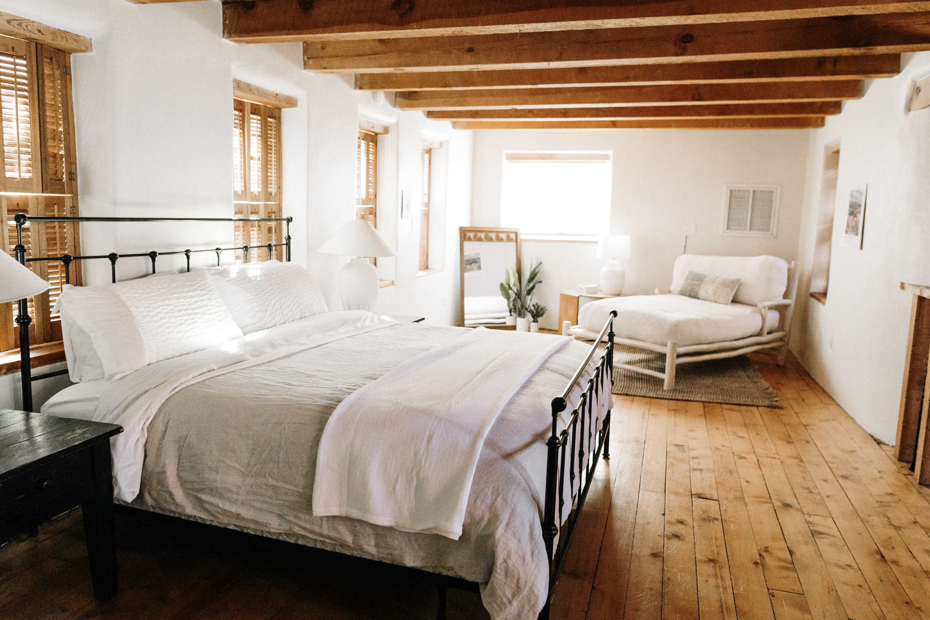 A spacious bedroom features a black metal bed with white linens, wooden beams overhead, and a cozy seating area with natural light streaming through the windows.