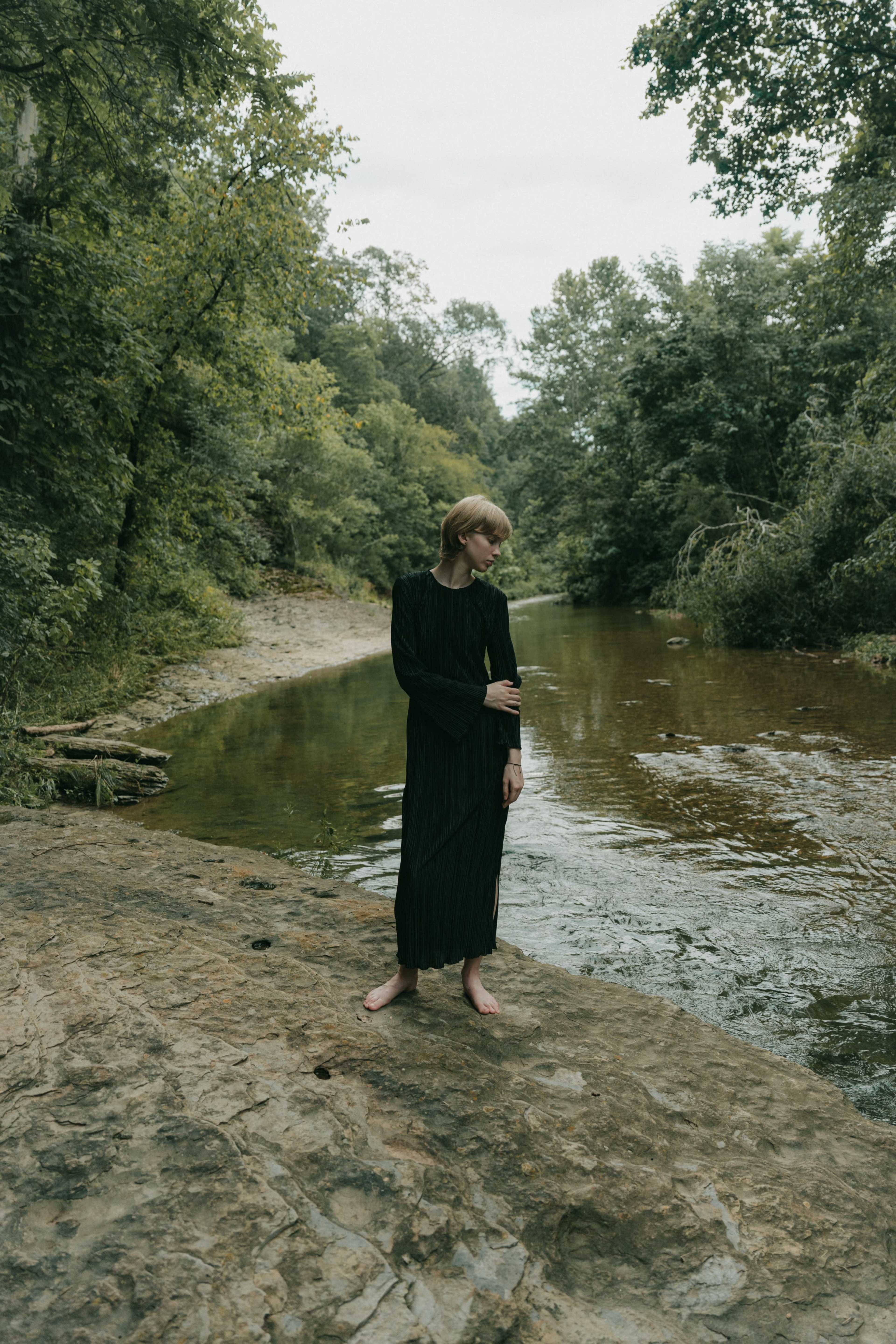 A person wearing a long black dress stands barefoot on a rocky bank beside a calm, tree-lined stream.