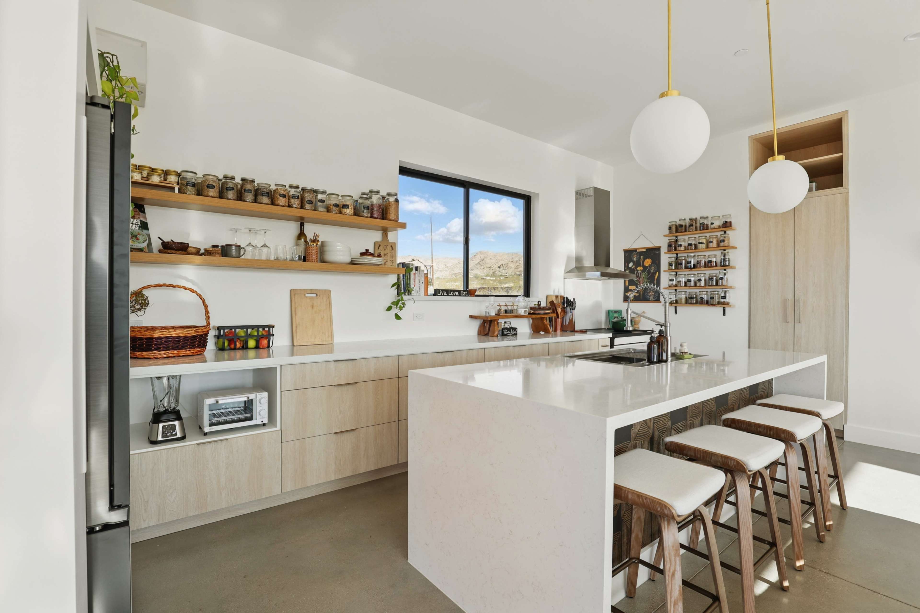 The kitchen features a minimalist design with a large island, wooden shelves holding various jars and utensils, and modern appliances.