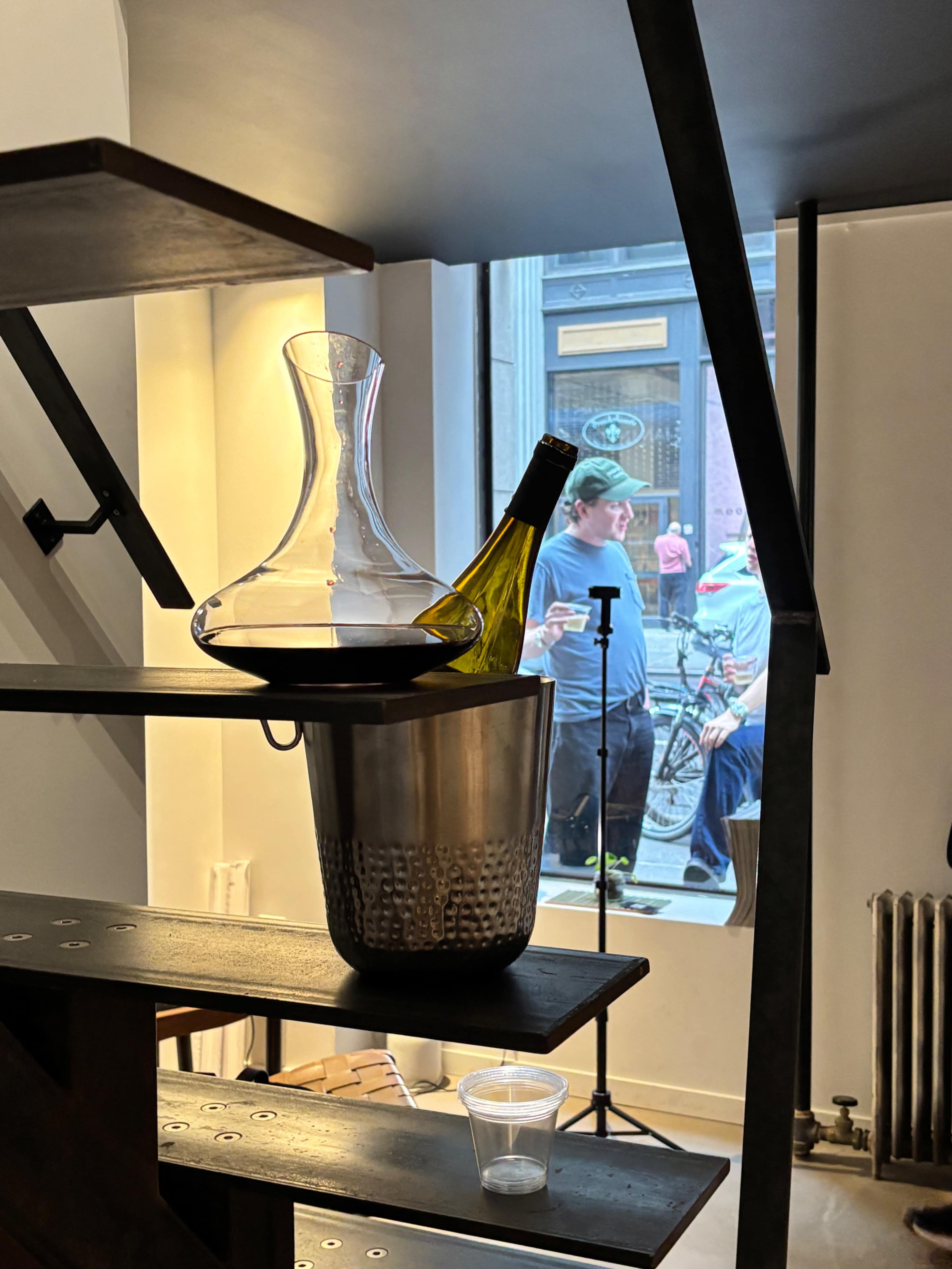 A wine decanter and a bottle of white wine rest in a bucket on a wooden shelf, while people can be seen outside through a window.