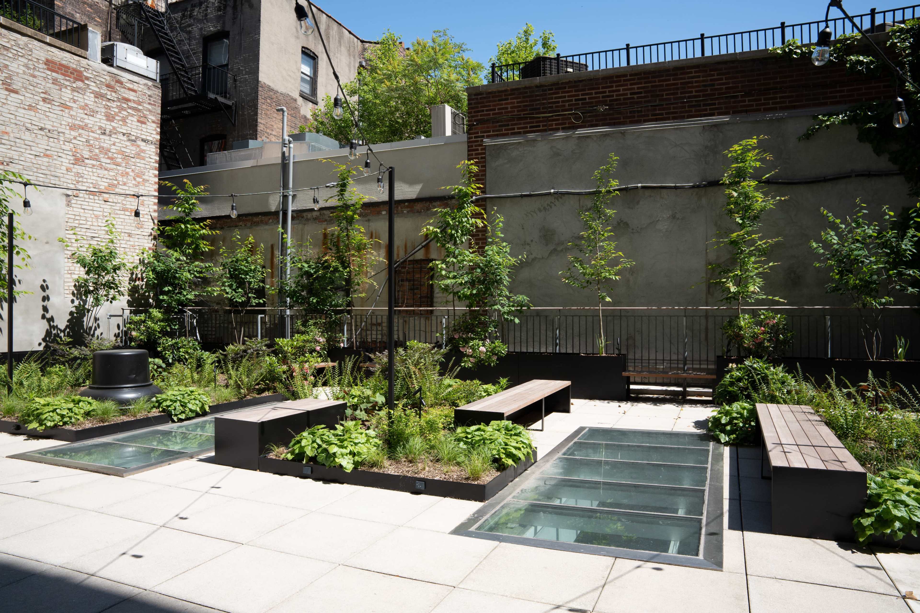The image shows a landscaped courtyard with several benches, glass panels, and greenery surrounded by buildings.