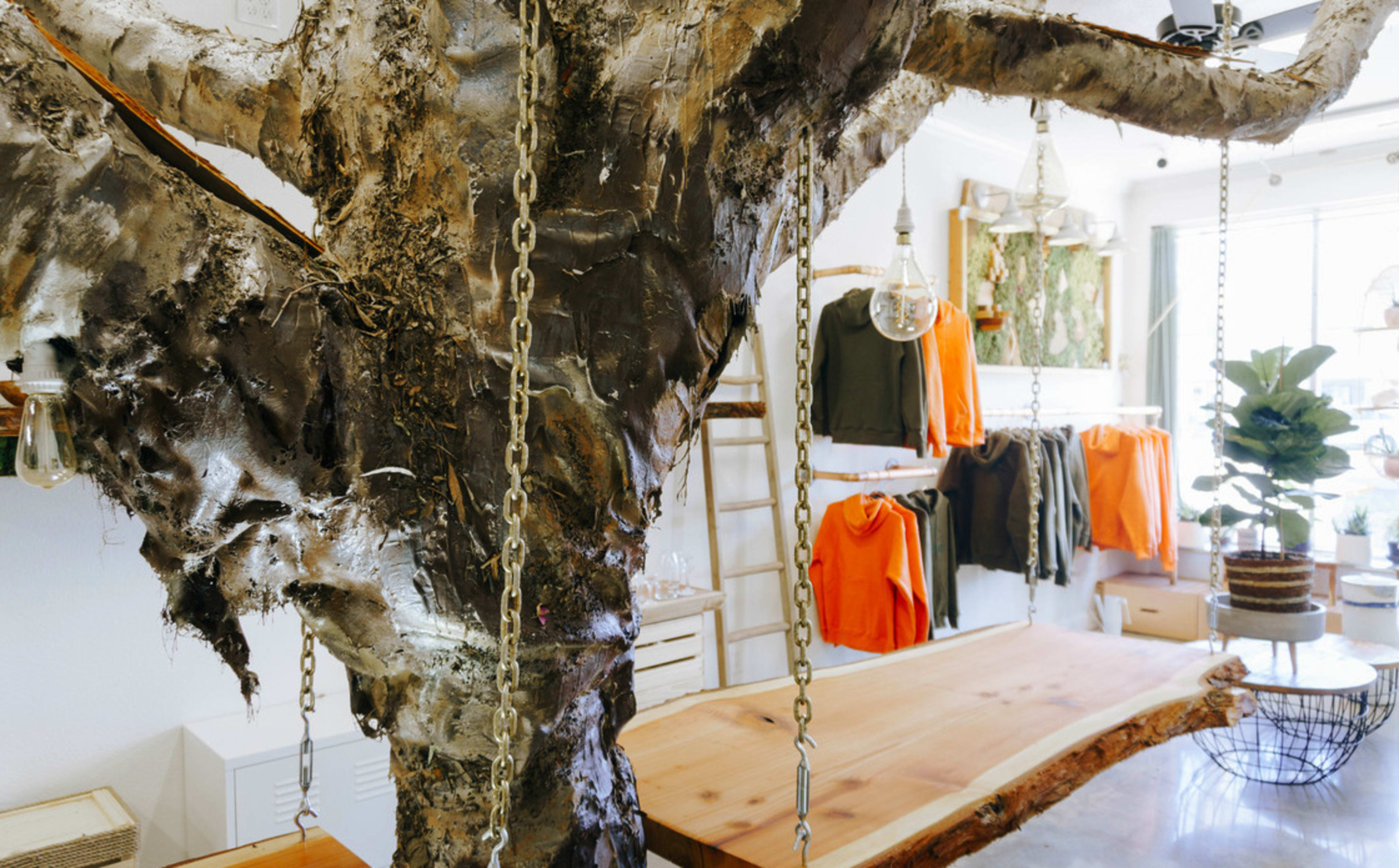 A large tree trunk is integrated into a retail space, adorned with swings and surrounded by hanging clothing items and decorative plants.