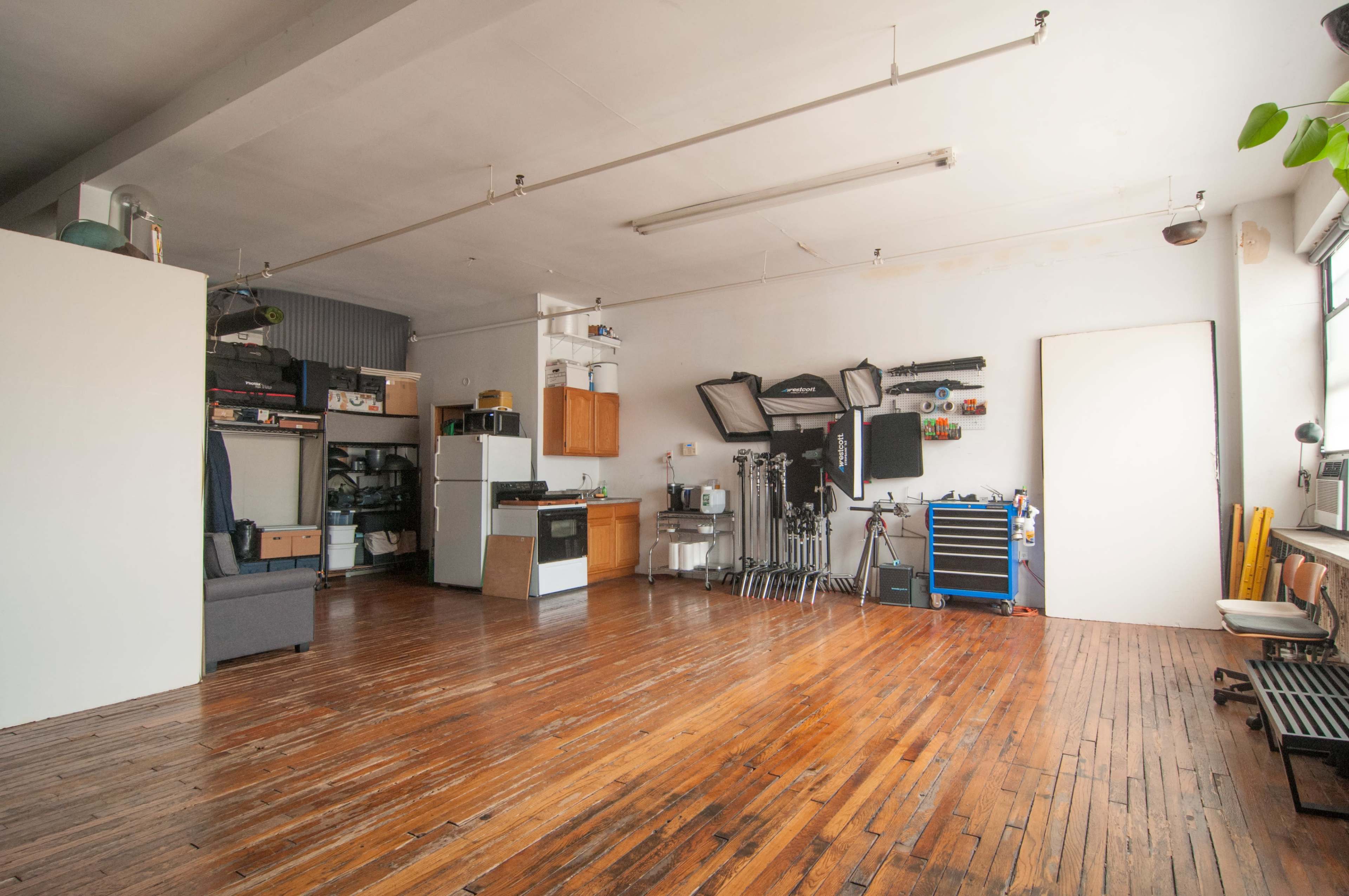 The image shows a spacious room with wooden flooring, featuring a kitchen area on one side and various equipment and storage on the opposite side.