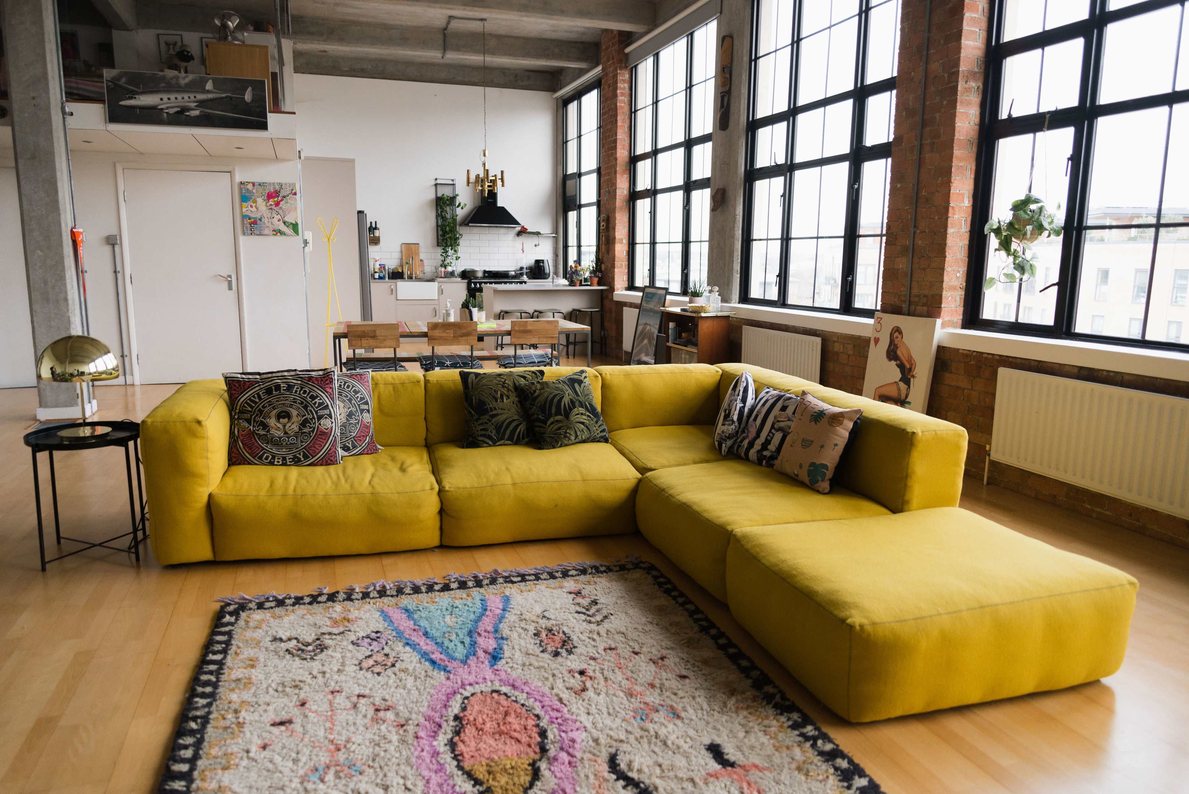 A yellow sectional sofa with decorative pillows is positioned in a spacious loft with large windows and an open kitchen in the background.