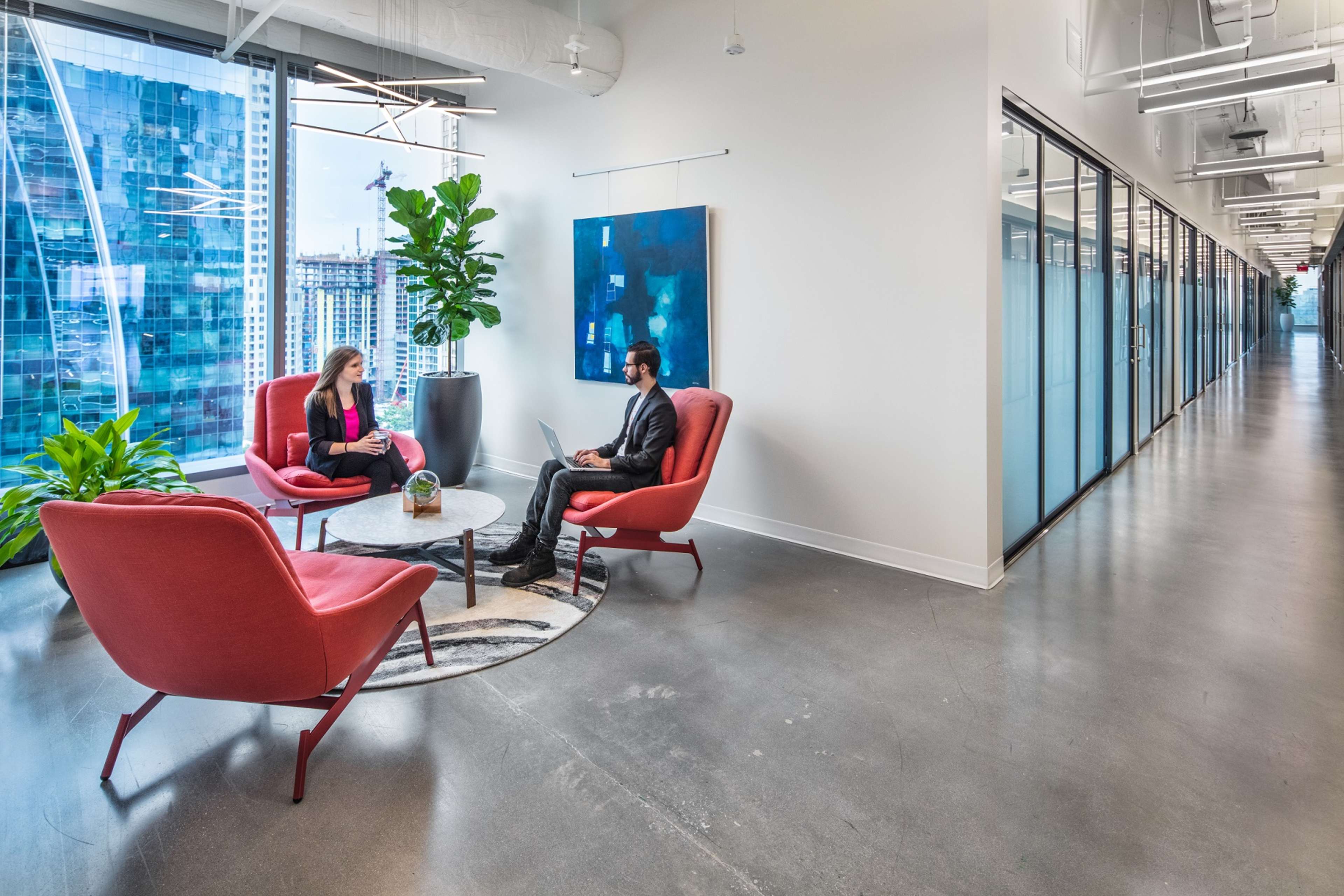 Two people sit in red chairs around a small table in a modern office space with large windows and a hallway lined with glass-walled offices.