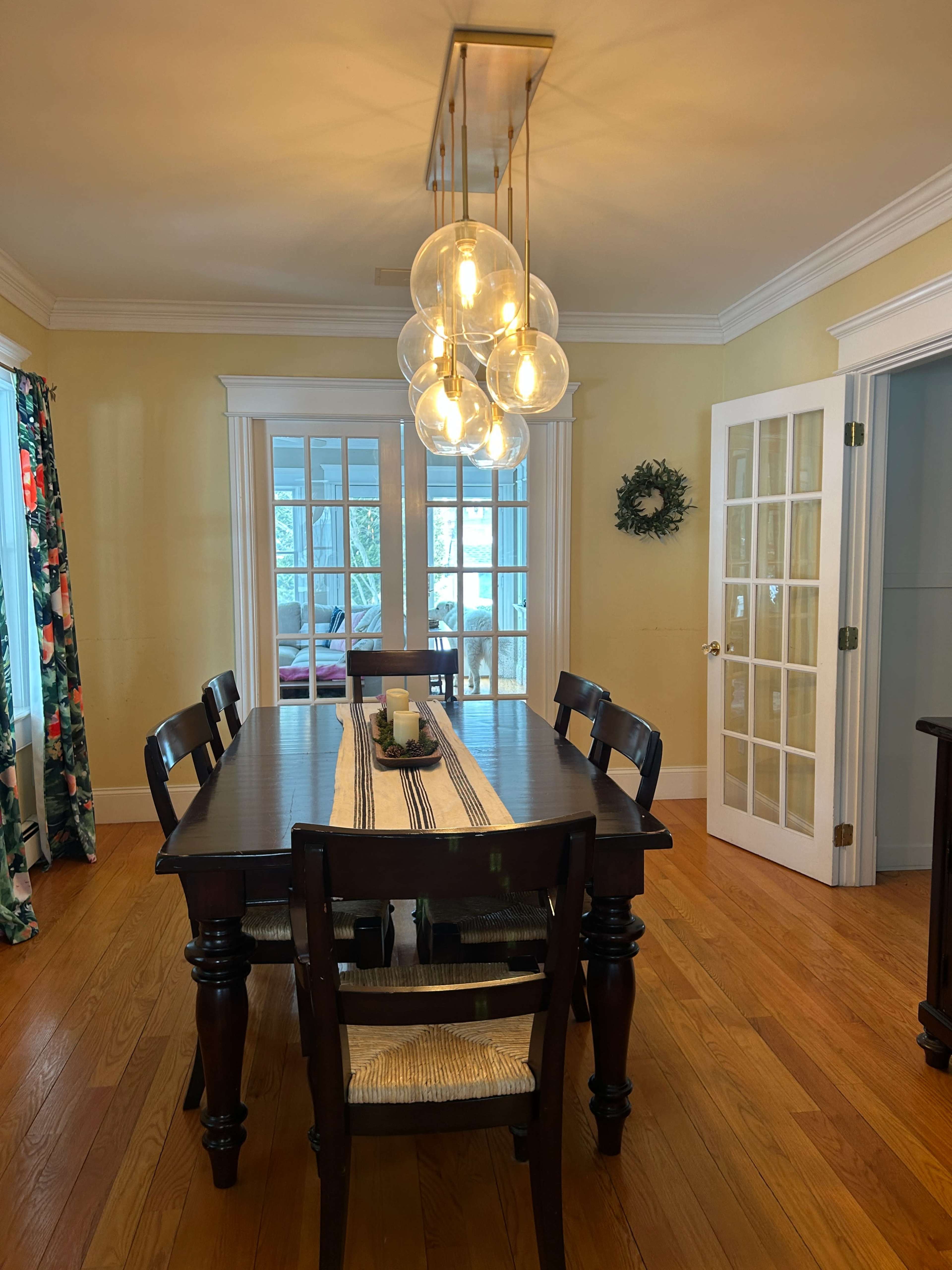 A wooden dining table with six chairs is illuminated by a modern light fixture in a room featuring yellow walls and French doors.