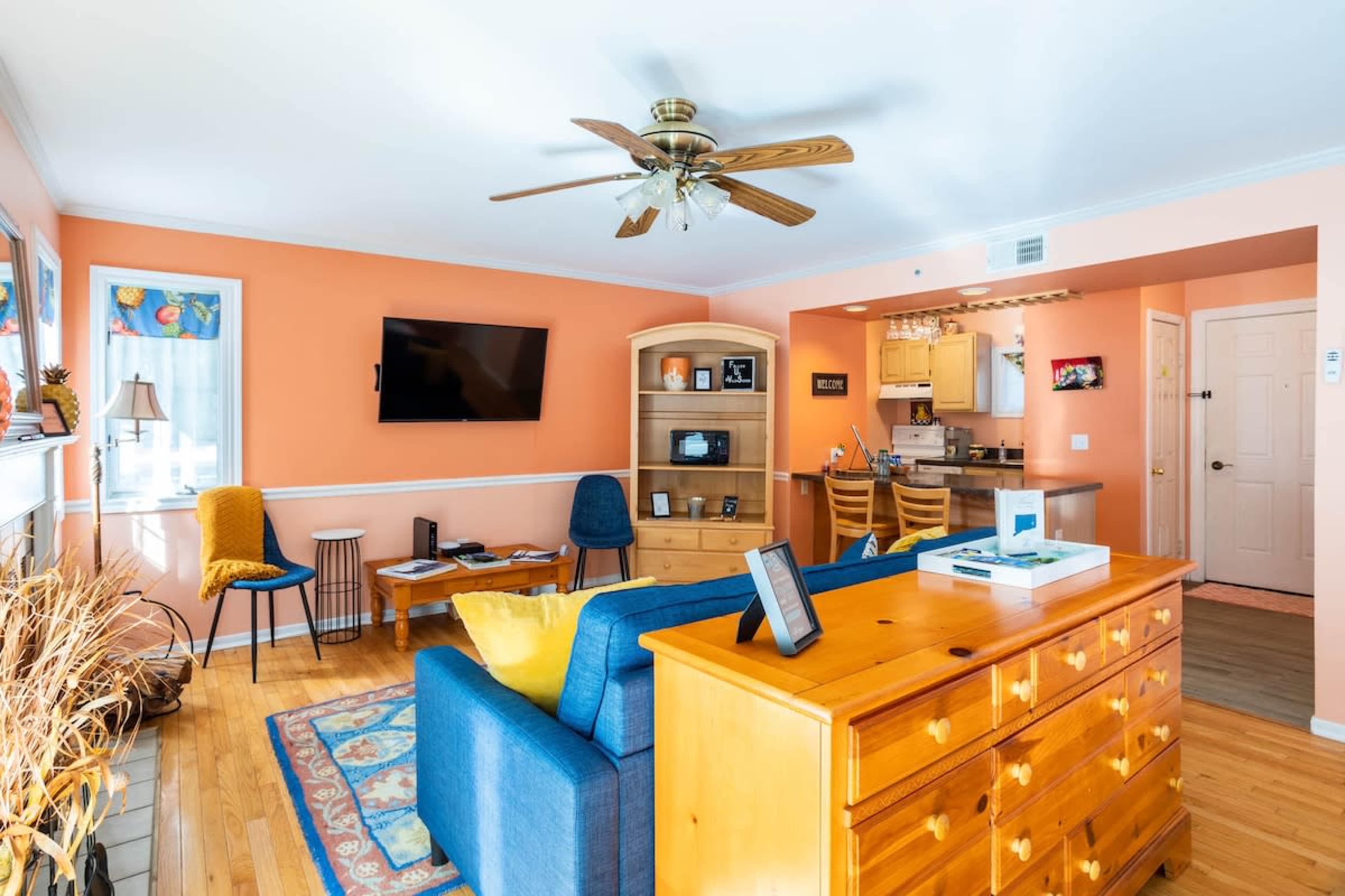 The image shows a bright living room with an orange wall, a blue sofa, wooden furniture, and a kitchen area visible in the background.