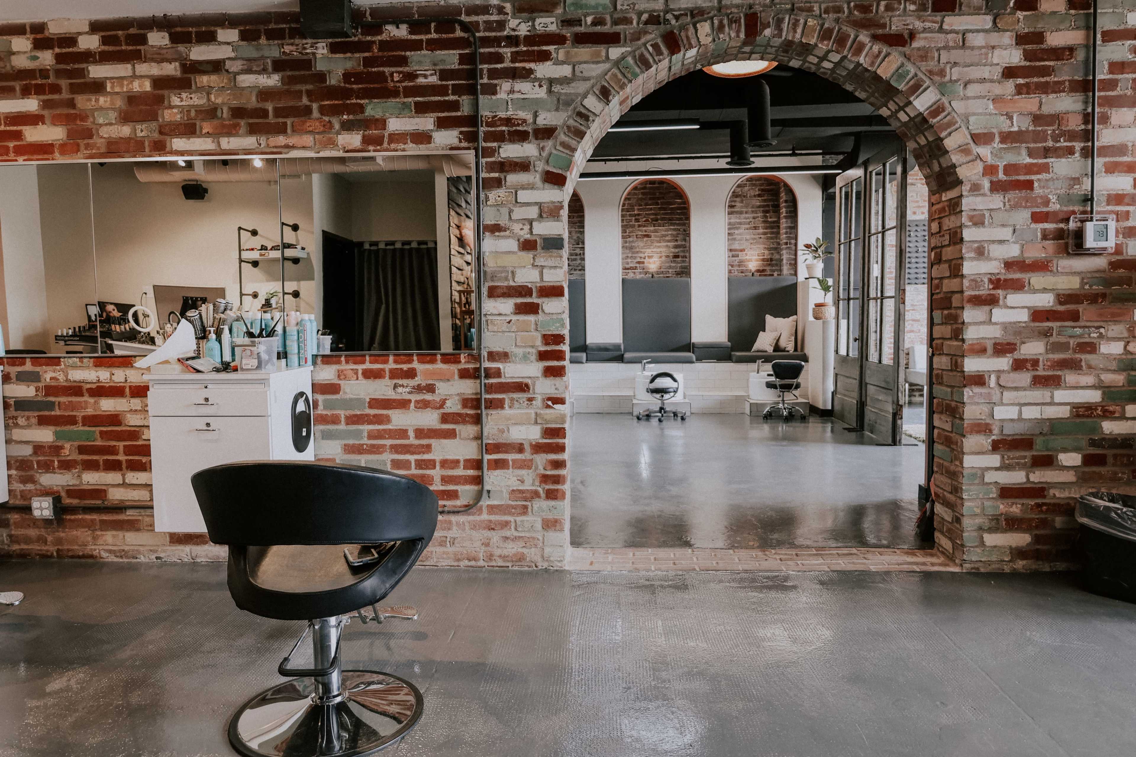 A hair salon with exposed brick walls, featuring a black stylist chair in the foreground and a spacious area with additional salon chairs in the background.