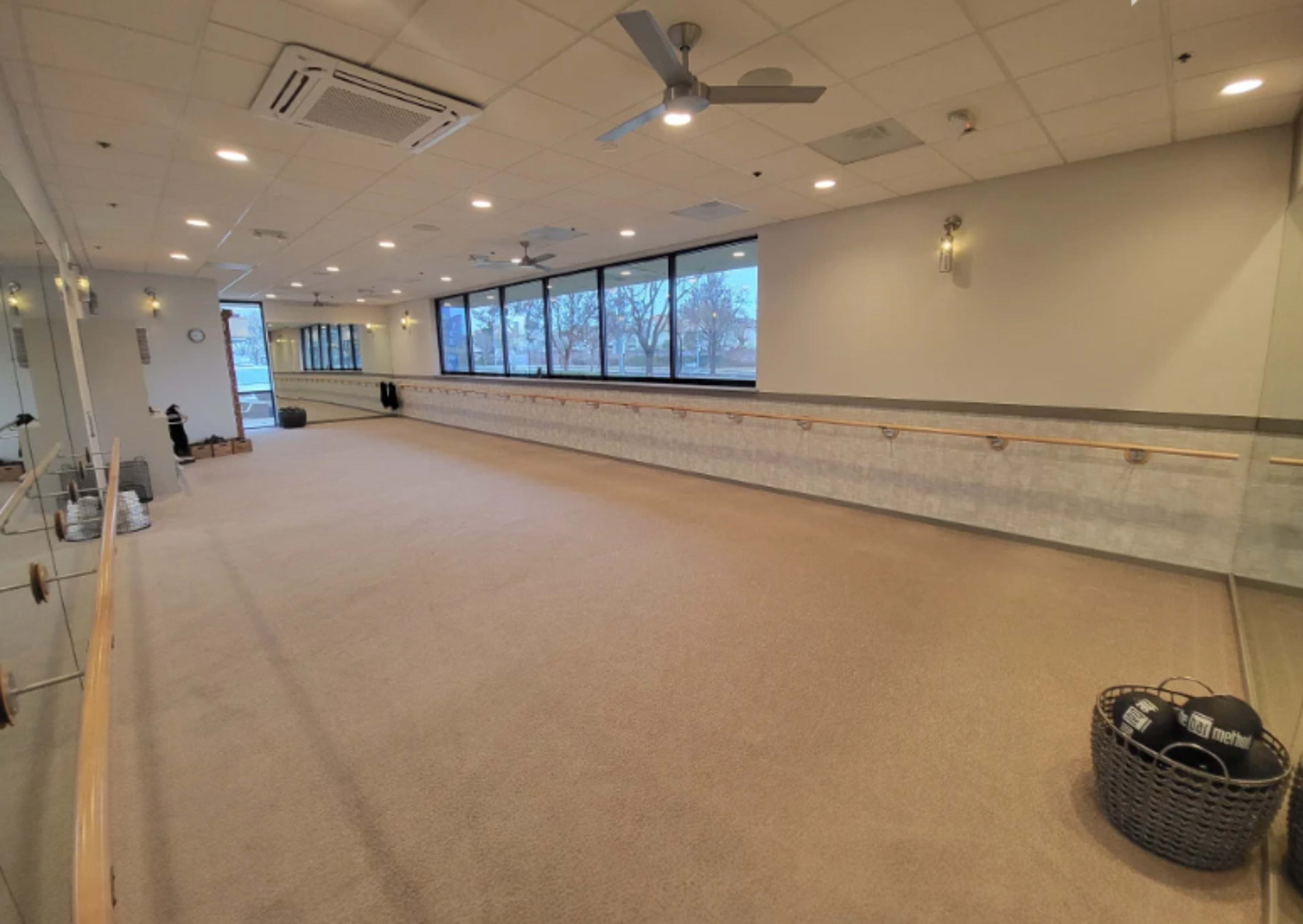 The image shows a spacious fitness studio with mirrored walls, a barre along one side, and large windows allowing natural light to fill the room.