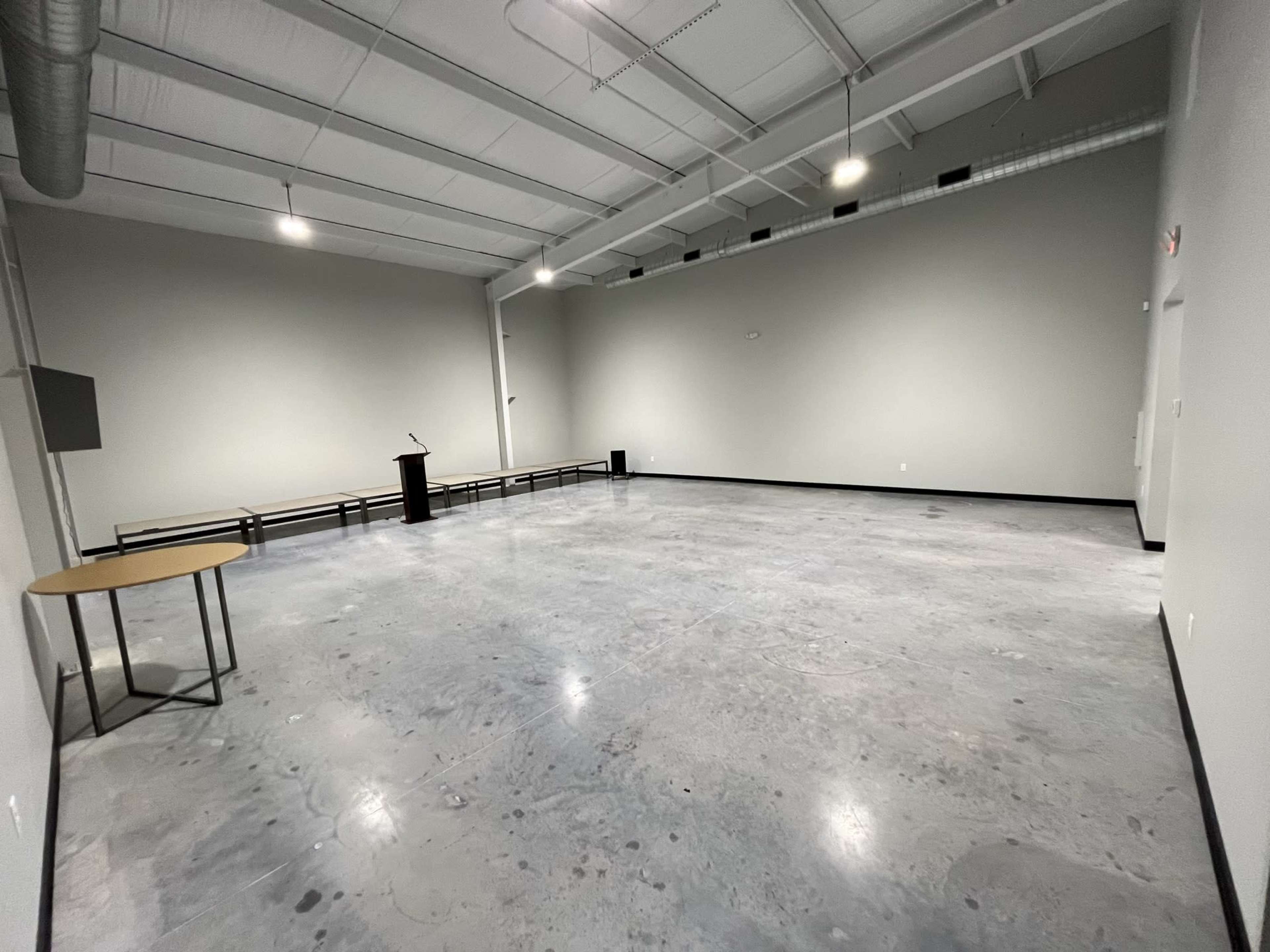 A spacious, empty room with a polished concrete floor, a podium at one end, and a table against the wall.