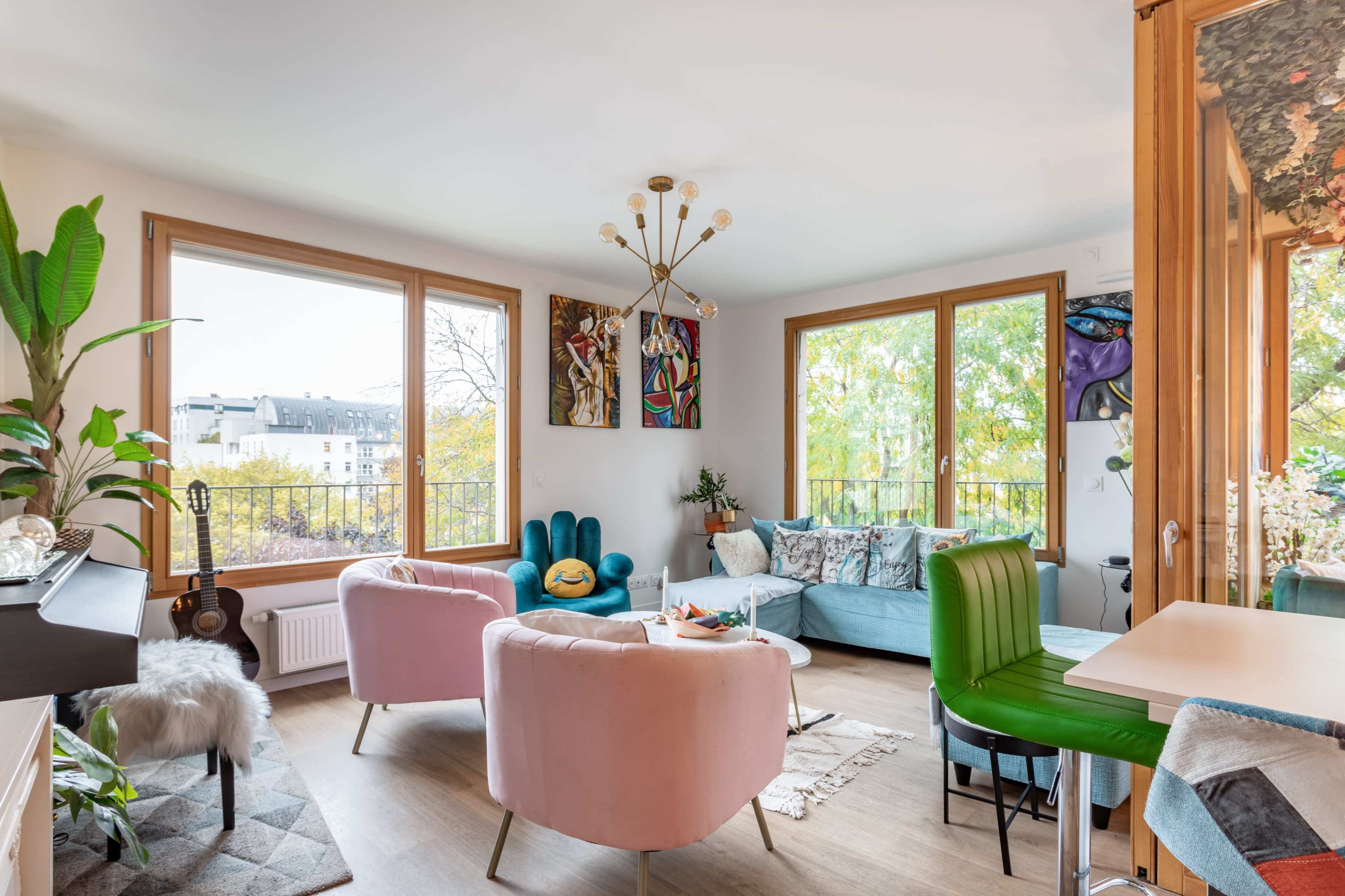 The living room features large windows, a mix of colorful furniture including teal and pink chairs, and decorative artwork on the walls.