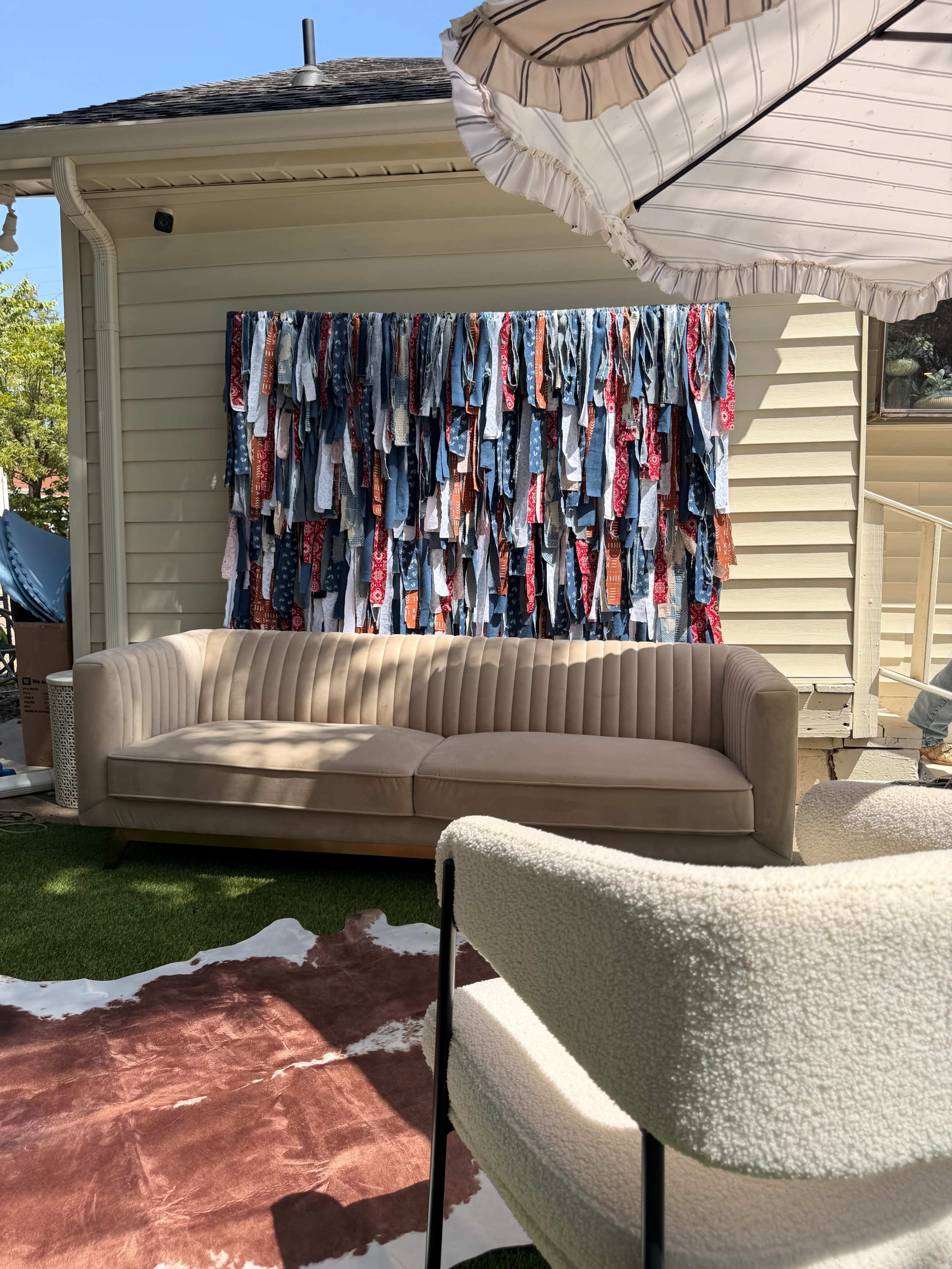 A beige couch and a plush chair are positioned in an outdoor area, with a wall covered in colorful pieces of fabric behind them, alongside a shaded umbrella.
