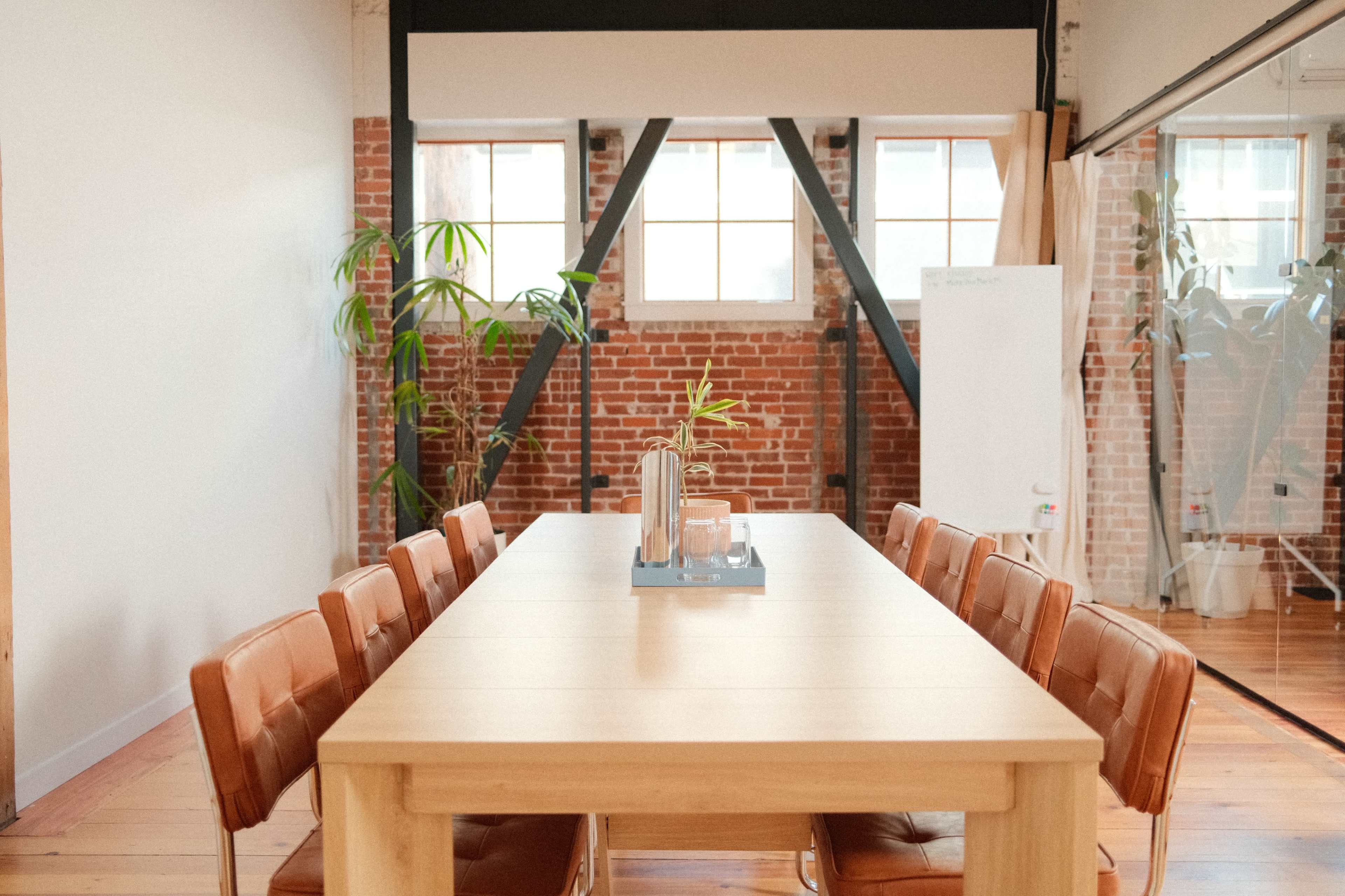 A long wooden conference table with brown leather chairs is positioned in a bright space featuring brick walls and large windows.