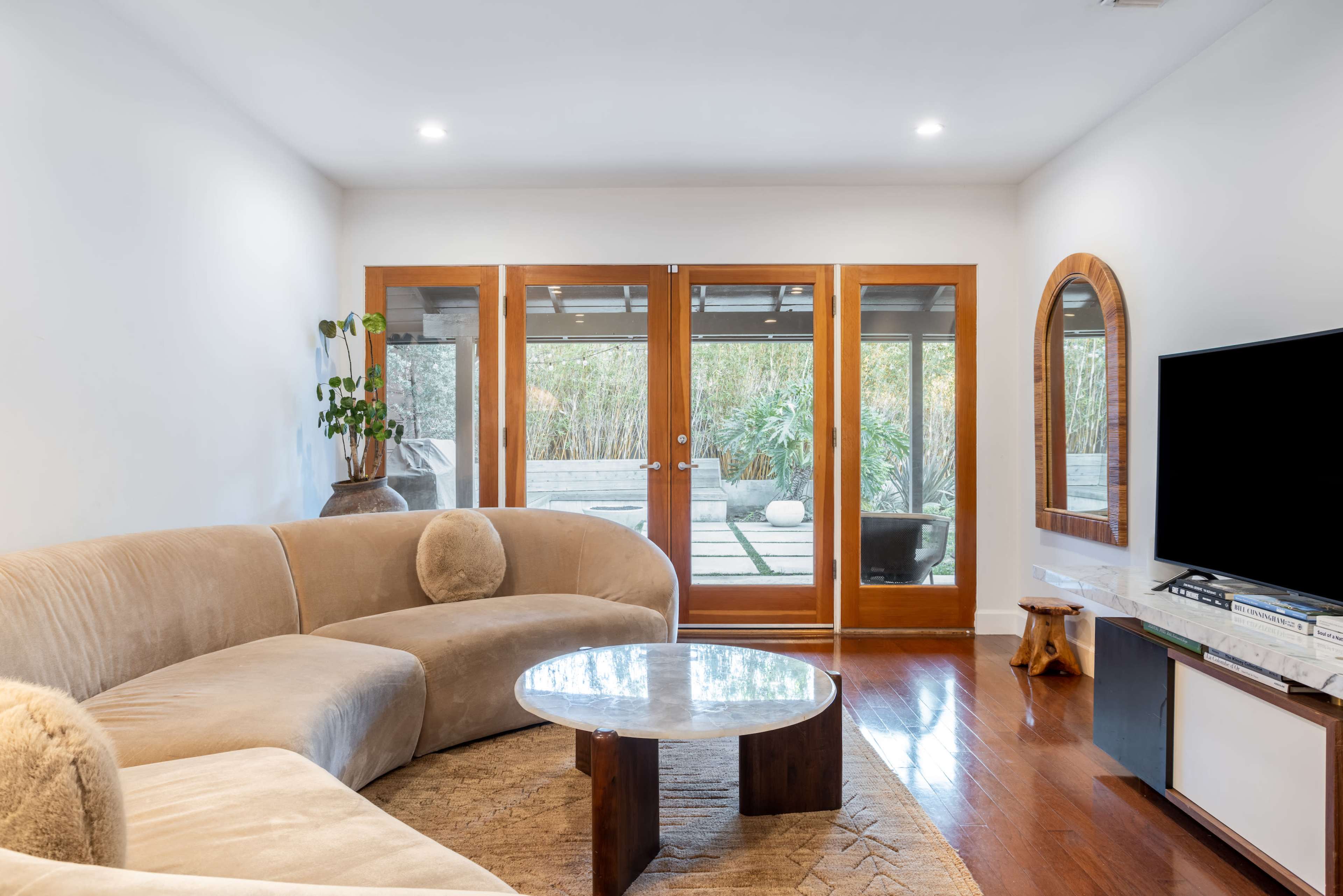 A modern living room features a curved beige sofa, a circular marble coffee table, and large glass doors opening to an outdoor garden.