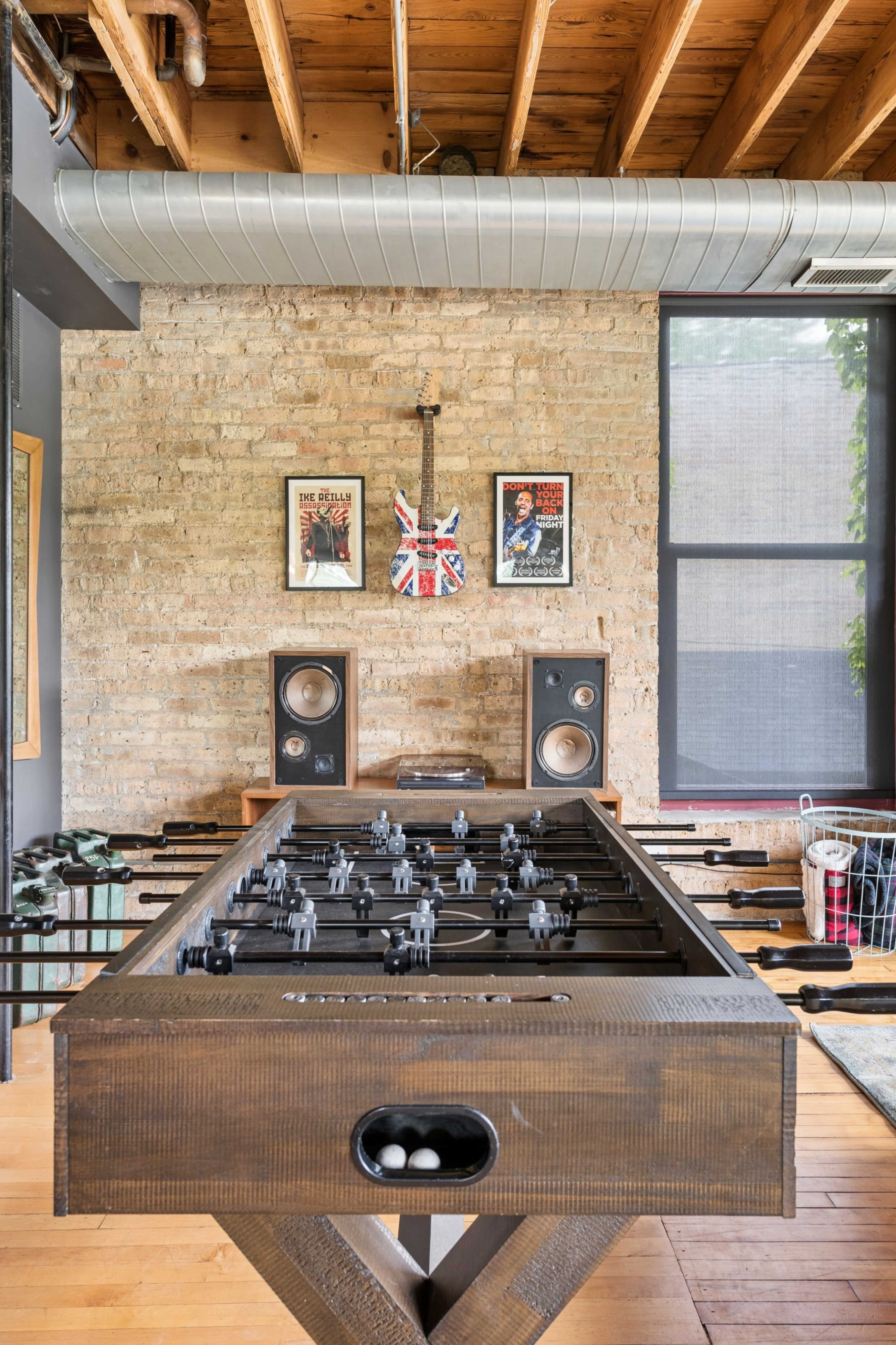 The image features a foosball table in a room with exposed brick walls, wooden beams, and framed posters, alongside speakers and a laundry basket.
