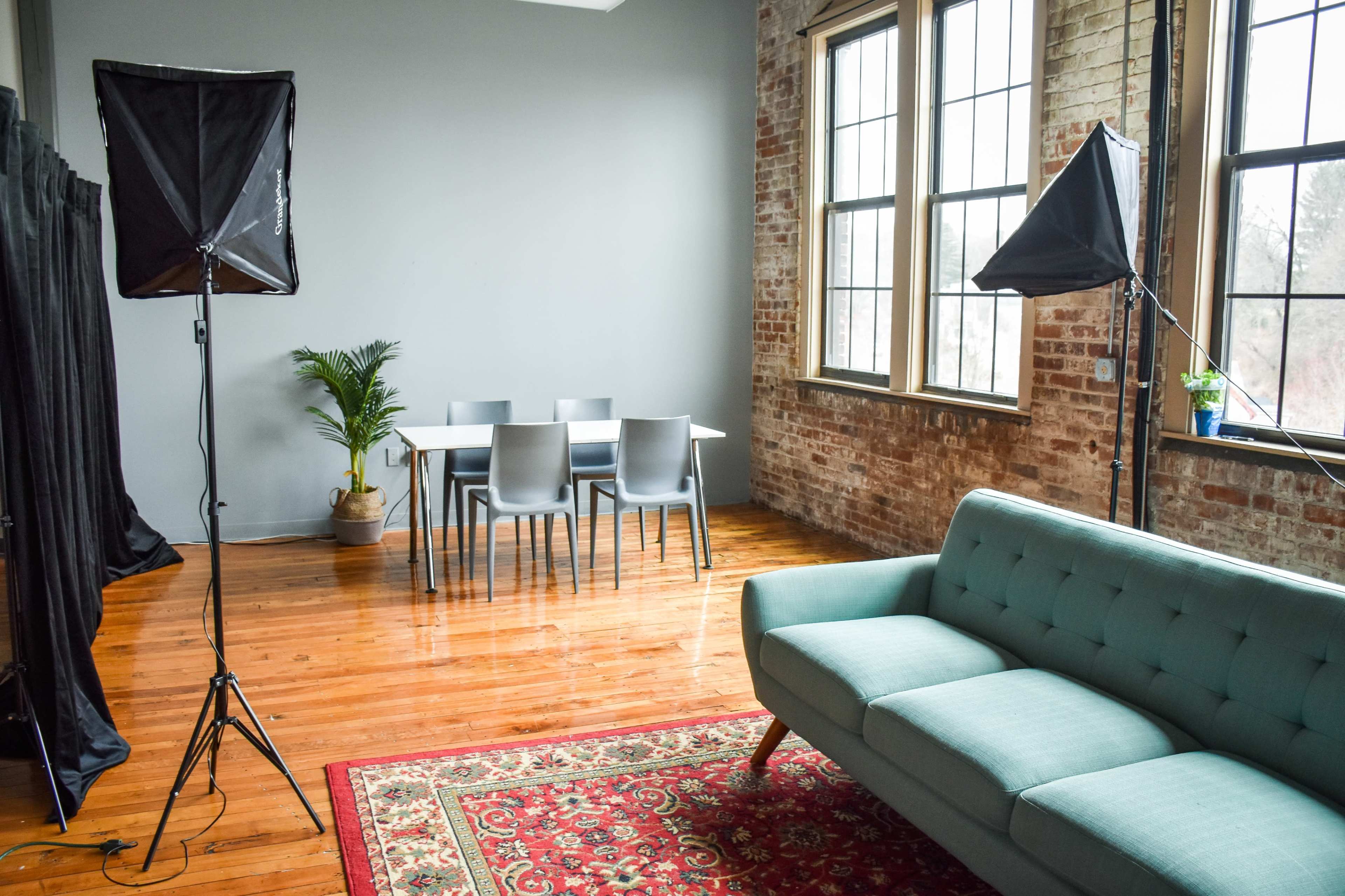 A bright room with a table and chairs in the center, flanked by studio lights, a plant, and a couch on a patterned rug.