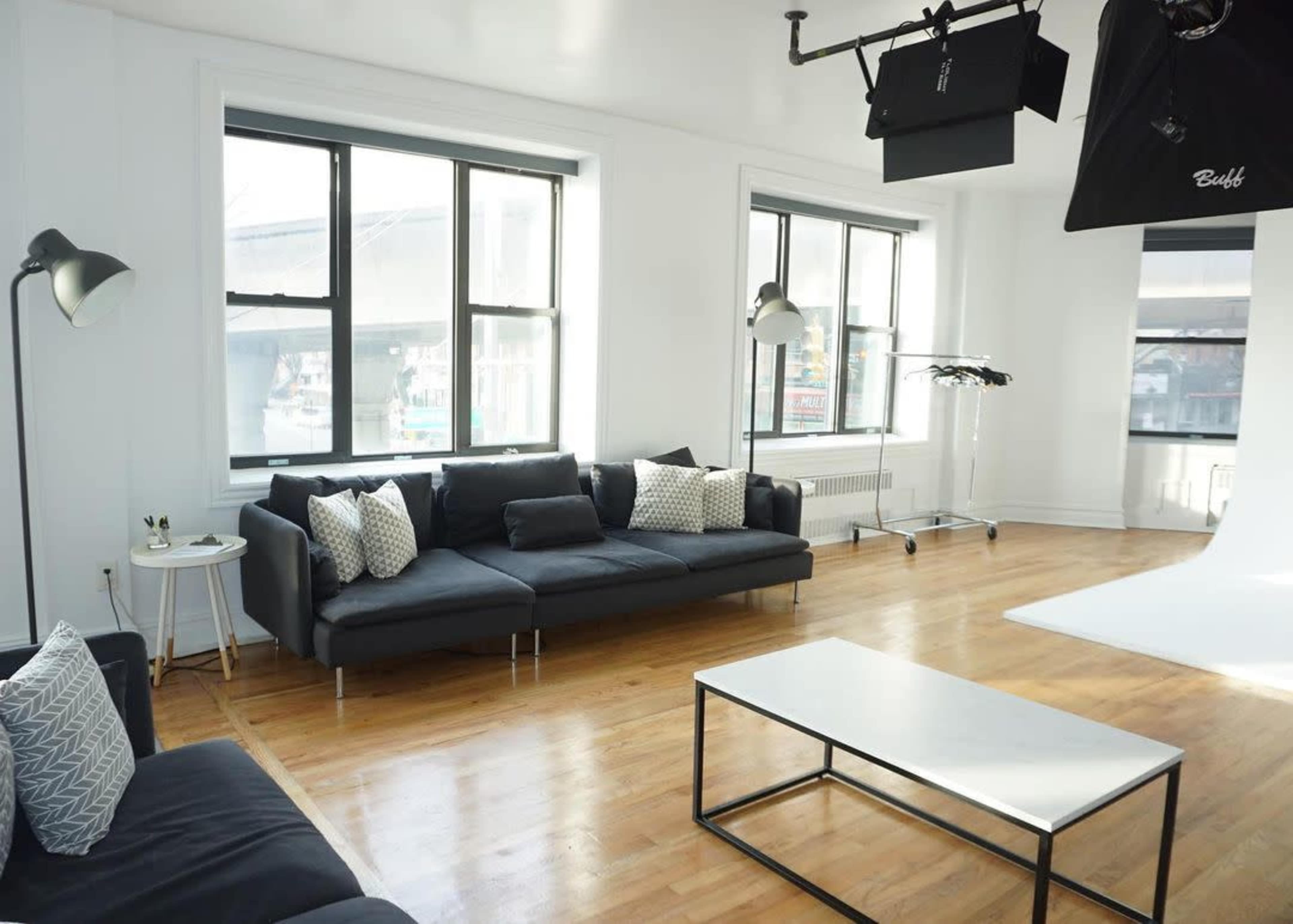 A bright, spacious room with large windows, a dark gray sectional sofa, a coffee table, and professional lighting equipment.