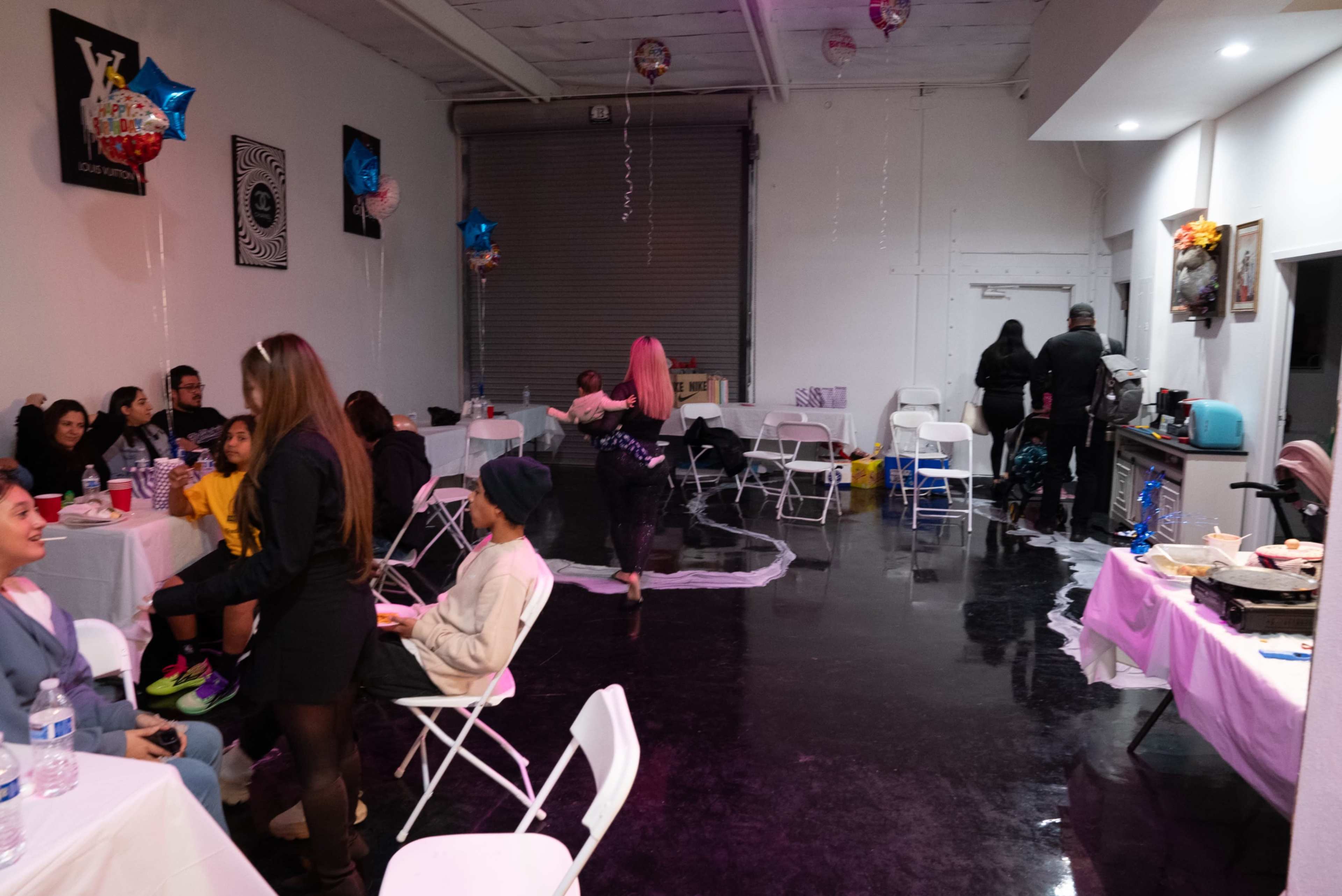 A group of people is gathered inside a spacious room decorated with balloons, with some sitting at tables while others move around in the background.