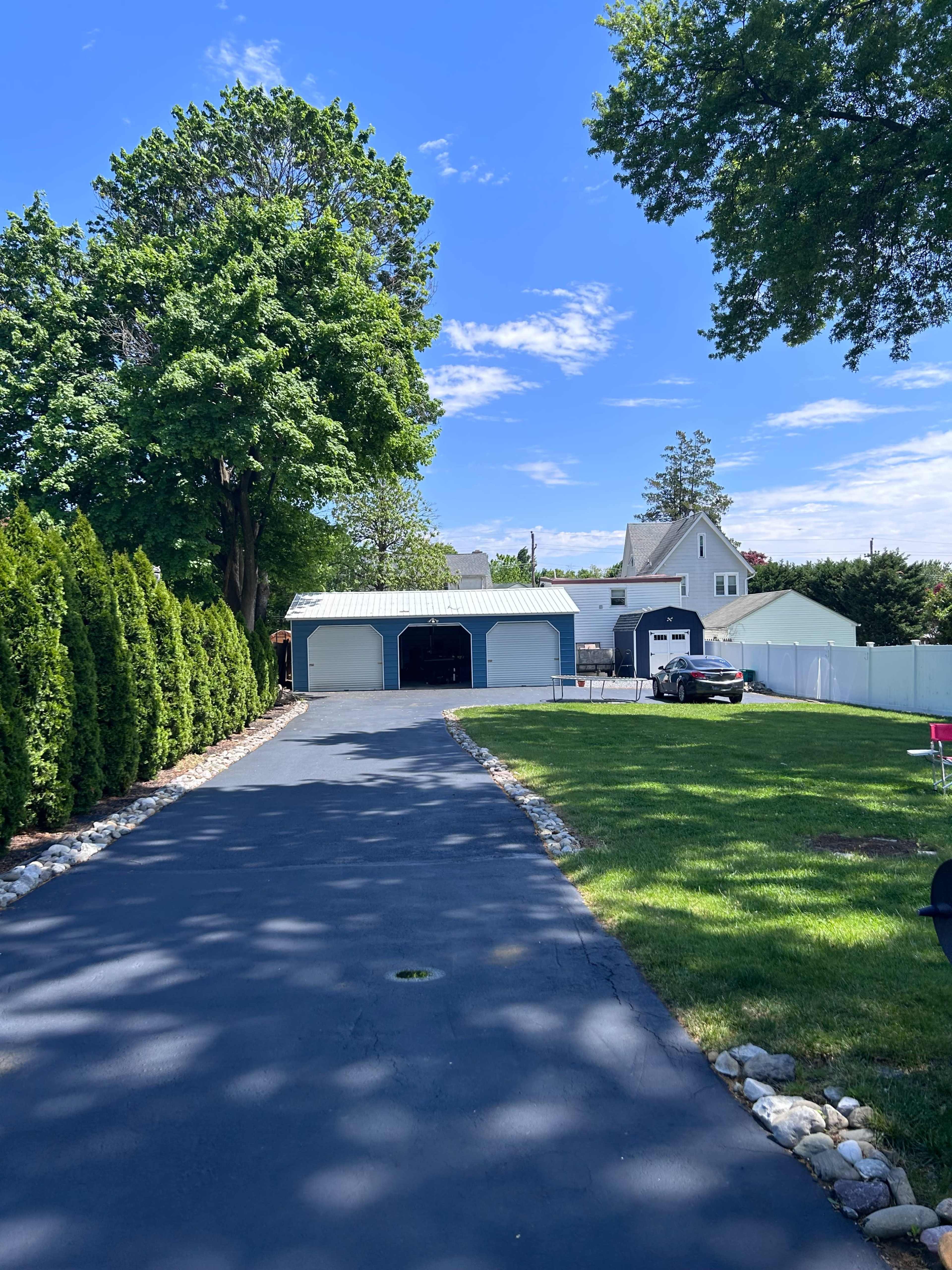 A paved driveway leads to a detached garage, with neatly trimmed hedges lining one side and a house visible in the background under a clear blue sky.