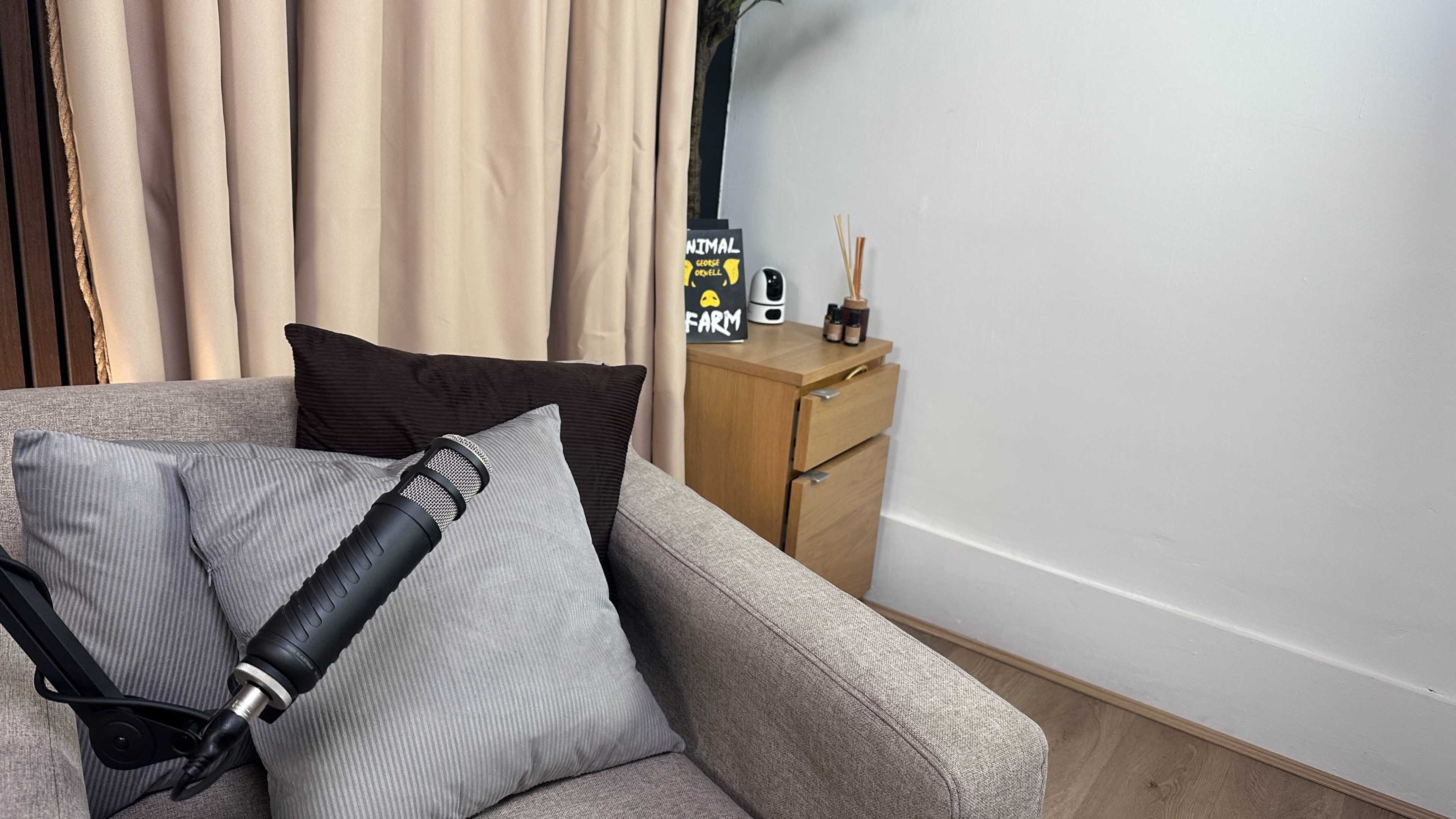 The image shows a corner of a room with a beige sofa, a microphone on the armrest, a small cabinet with an item on top, and a wall with curtains.