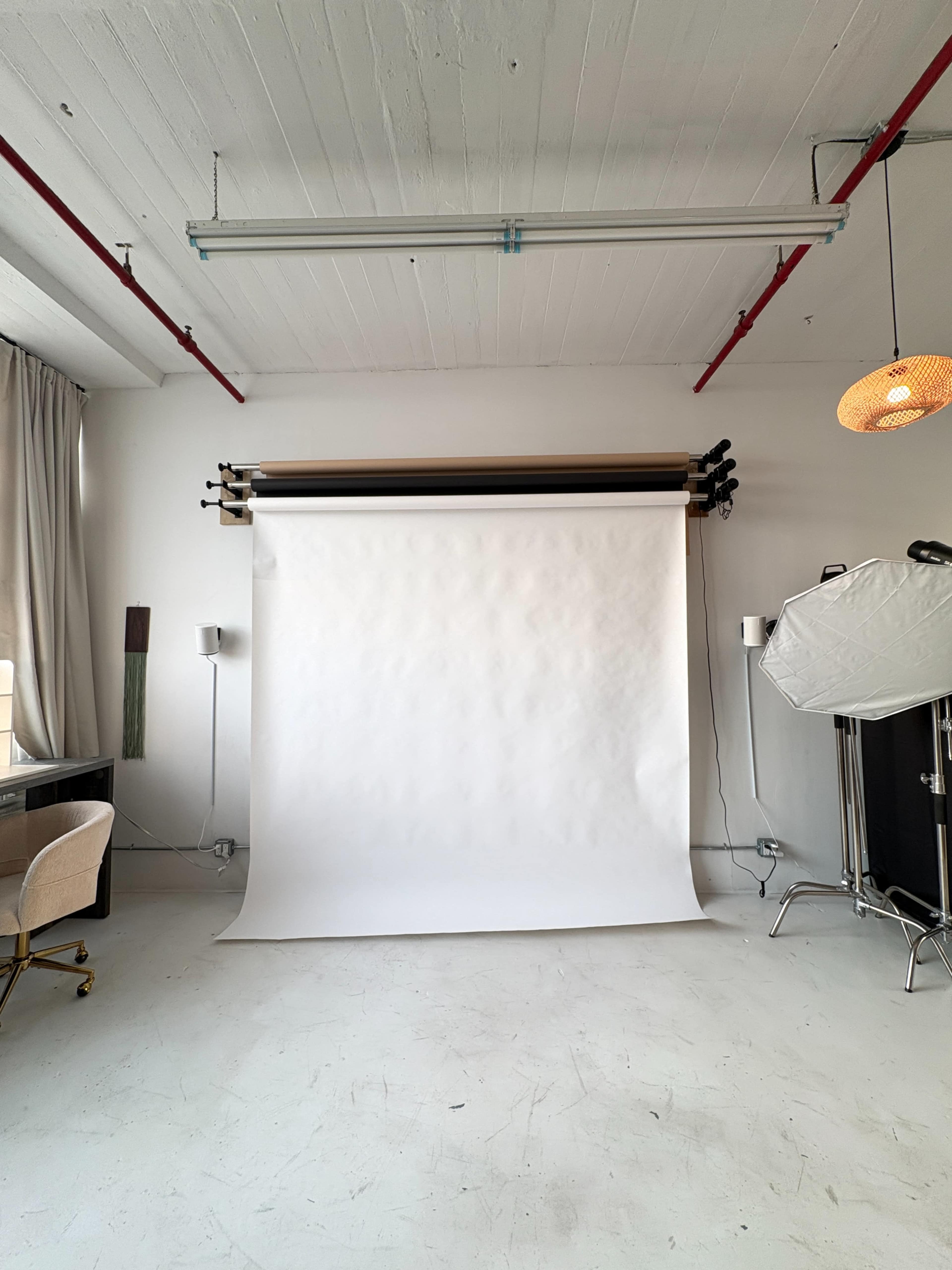 The image shows a bright, minimalist studio space featuring a white backdrop, a desk with a chair, and studio lighting equipment.